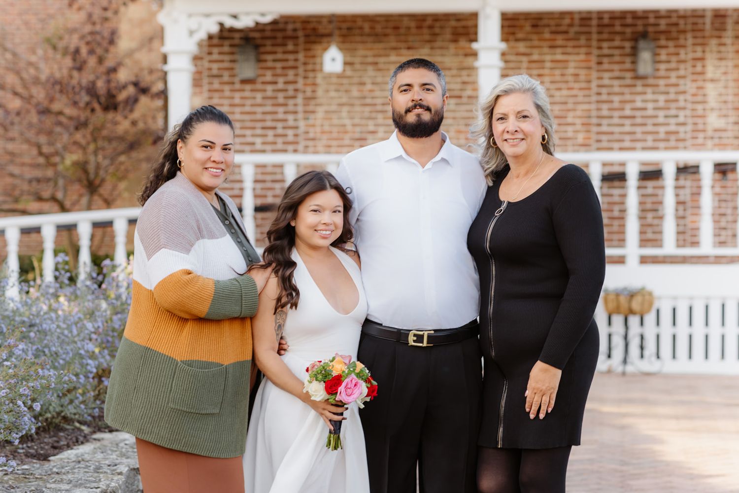 A bride and groom pose with two family members outside a brick building on their wedding day.