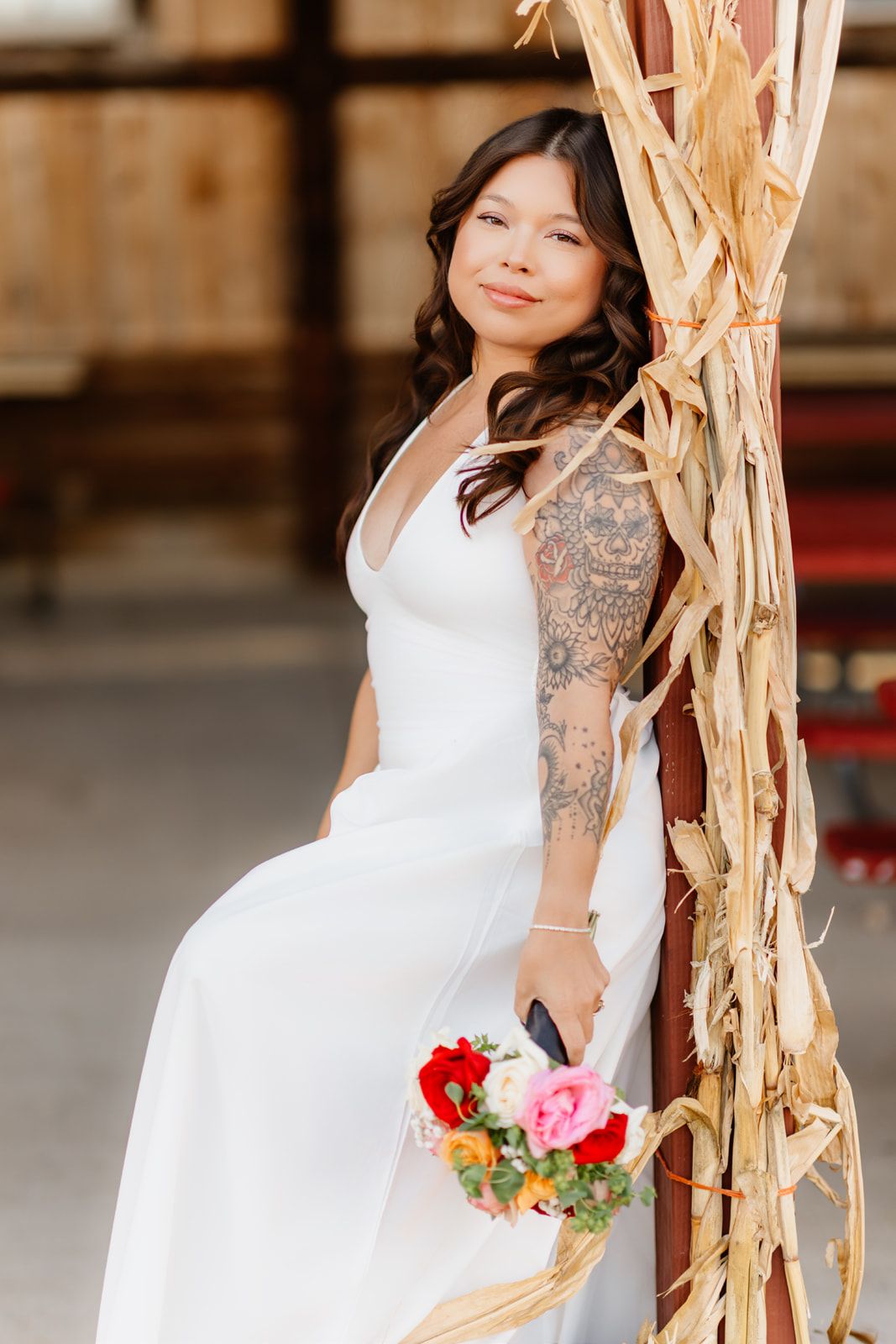Bride in white gown with arm tattoo holds colorful bouquet while leaning against rustic barn decor.