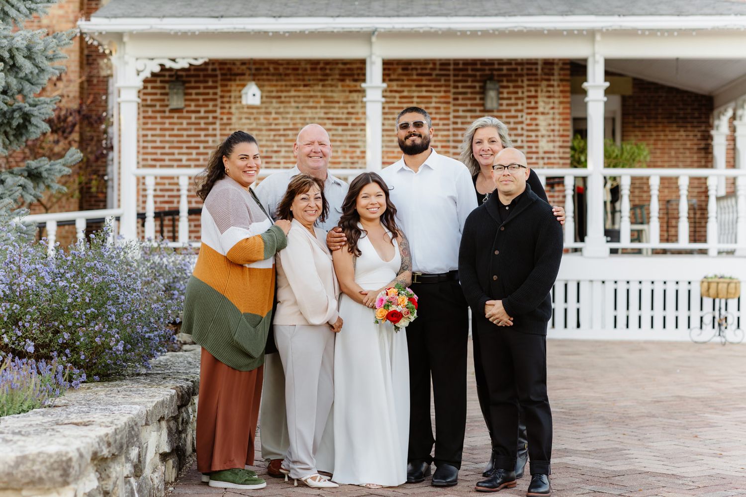 Wedding couple poses with family outside brick venue, bride in white gown holding colorful bouquet.