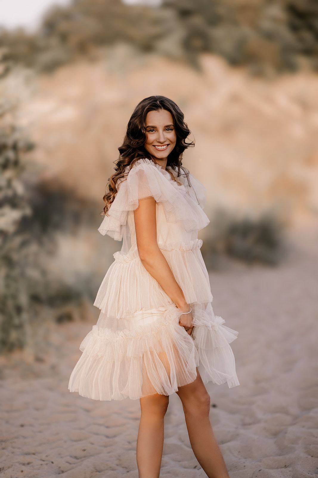 Smiling brunette woman in a ruffled beige tulle dress posing on a sandy path at golden hour.