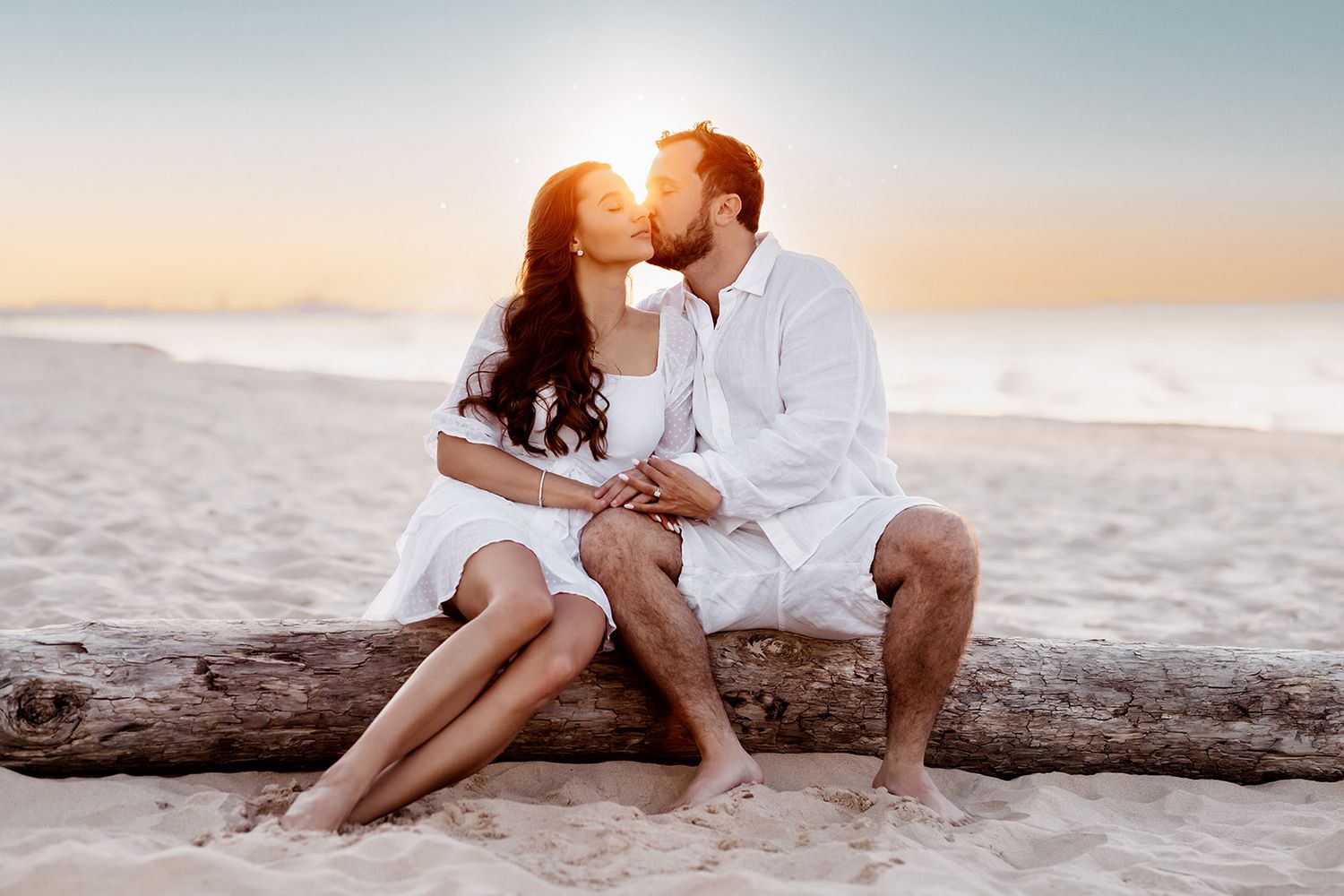 Couple sharing a kiss on driftwood at a sunny beach, both dressed in white outfits at sunset.