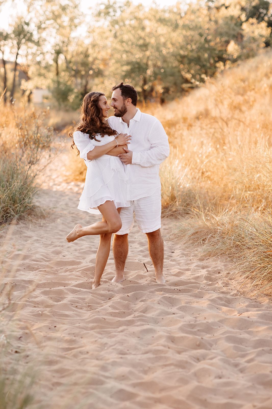 Couple in white outfits embracing on sandy beach path surrounded by golden grass in warm sunlight.
