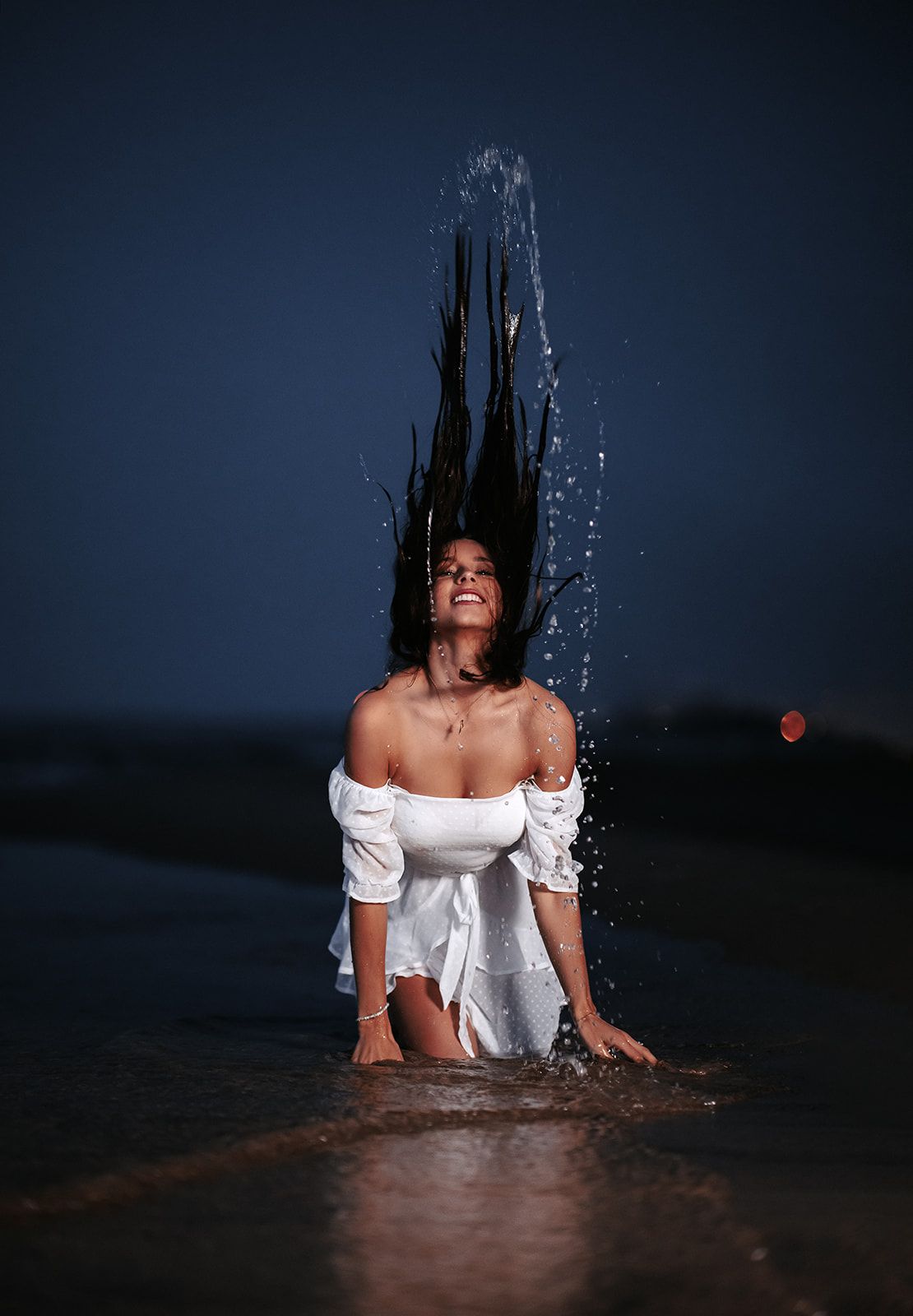 Woman in white off-shoulder dress flipping wet hair at beach shore during dusk, water droplets flying dramatically.
