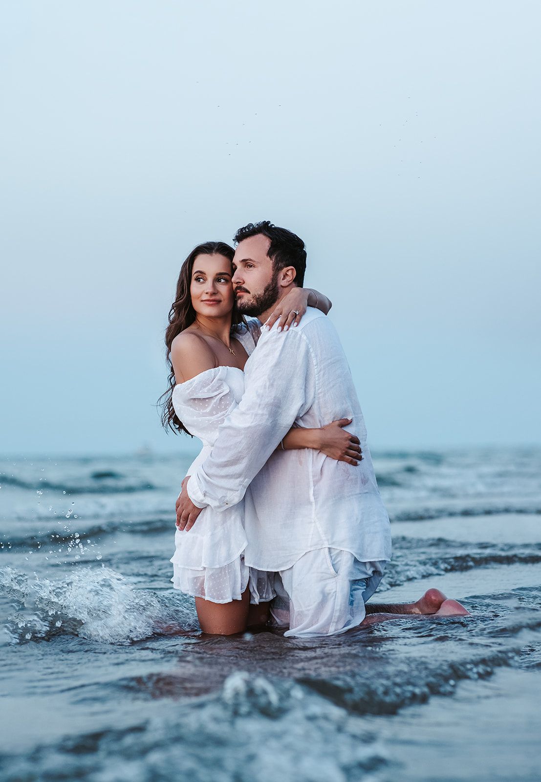 Romantic couple in white outfits embracing in ocean waves on a beach during a dreamy photoshoot.