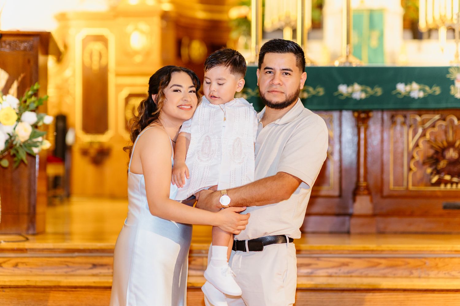 A couple smiles holding their child in white baptism attire inside a beautifully lit church.