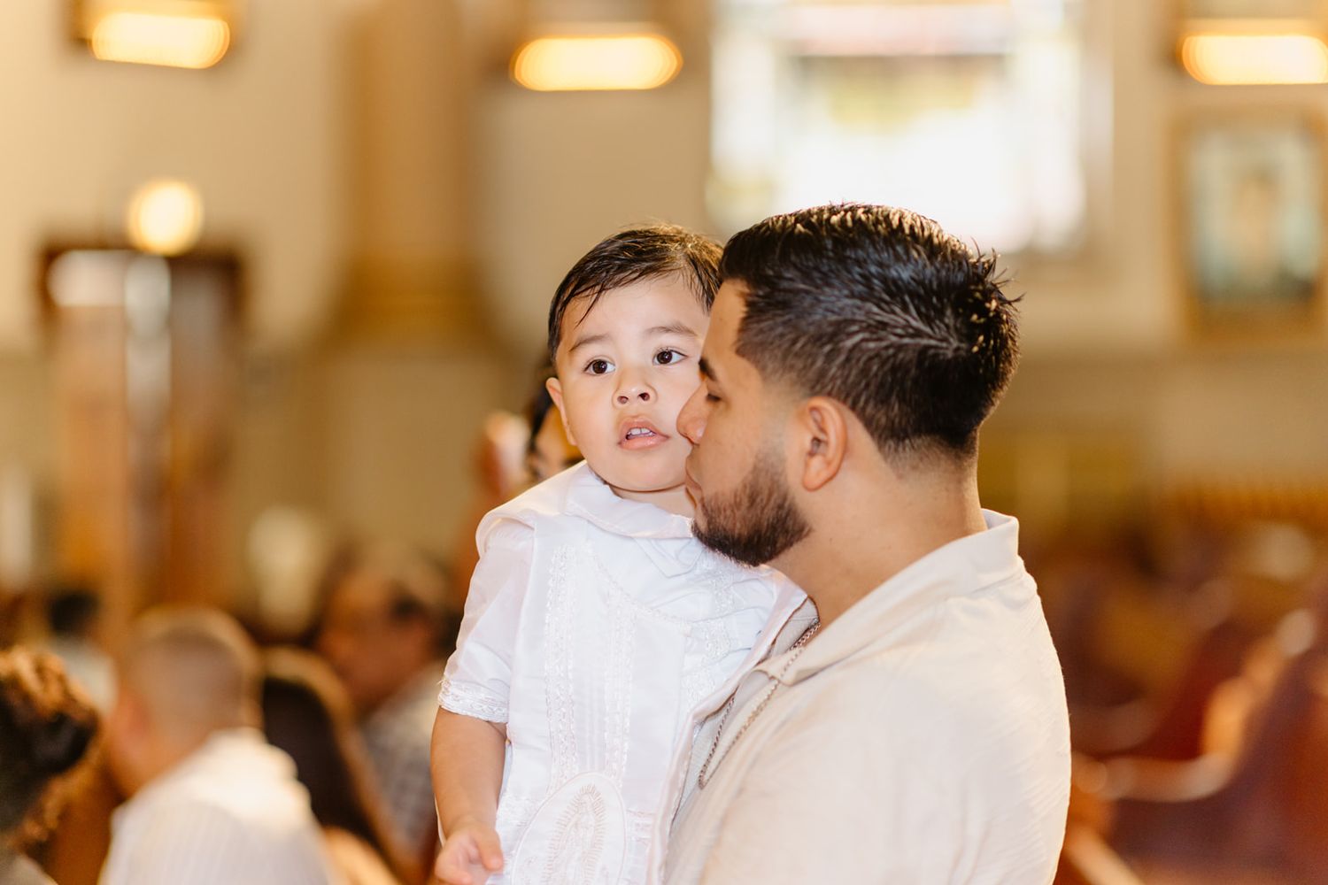 Father tenderly kisses his young child dressed in white during a baptism ceremony inside a church.