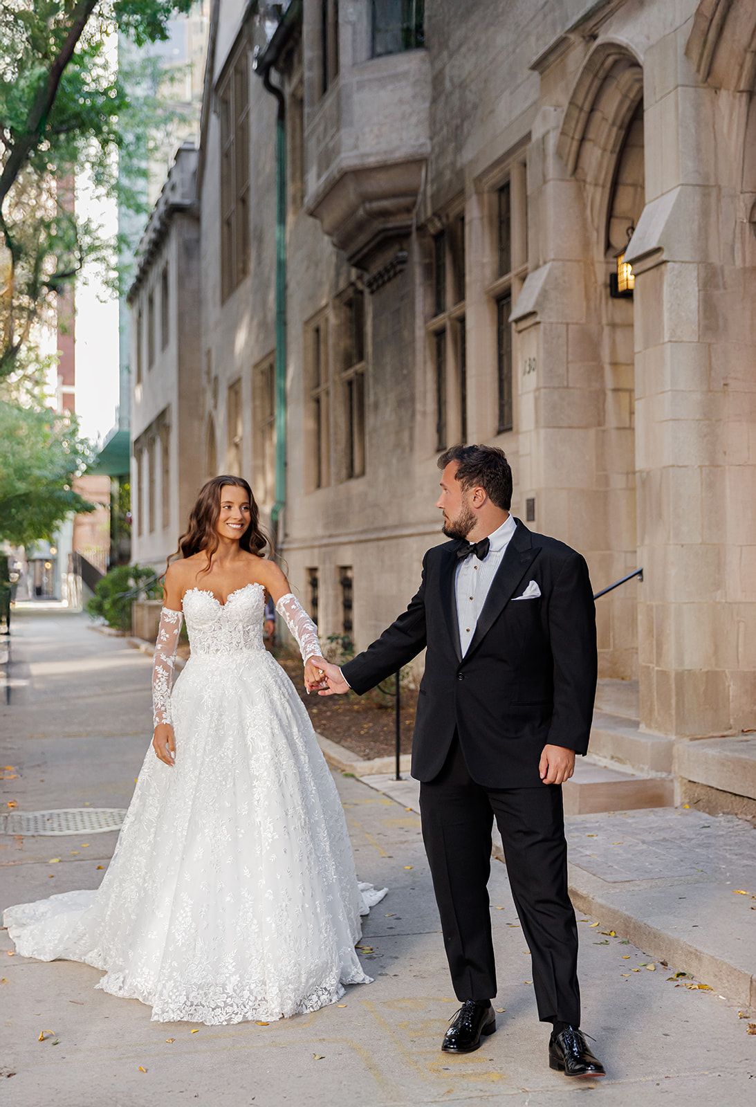 Couple in wedding attire hold hands walking on urban sidewalk beside elegant stone architecture.