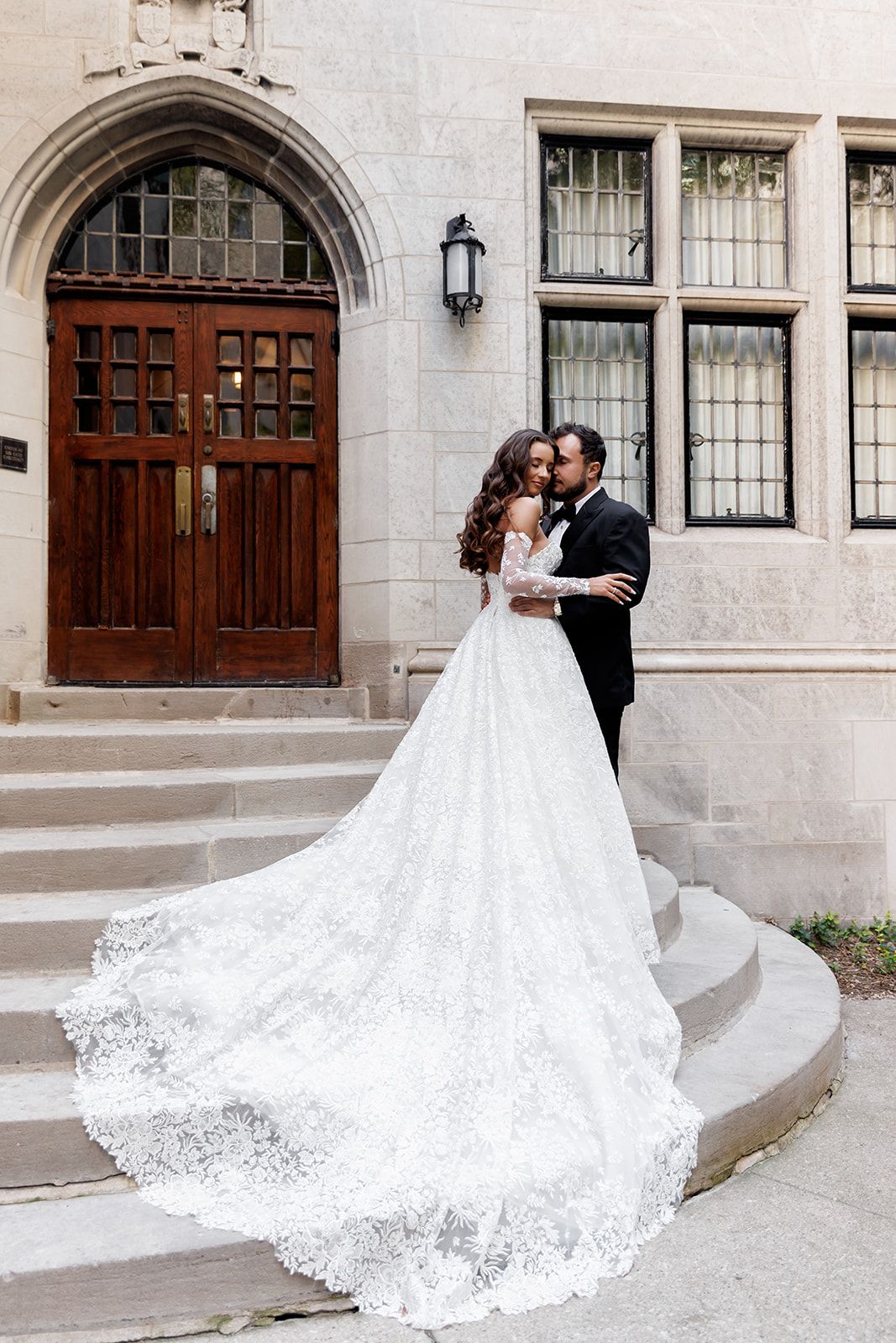 Bride in elegant lace ball gown with long train poses with groom in tuxedo on stone steps of historic building.