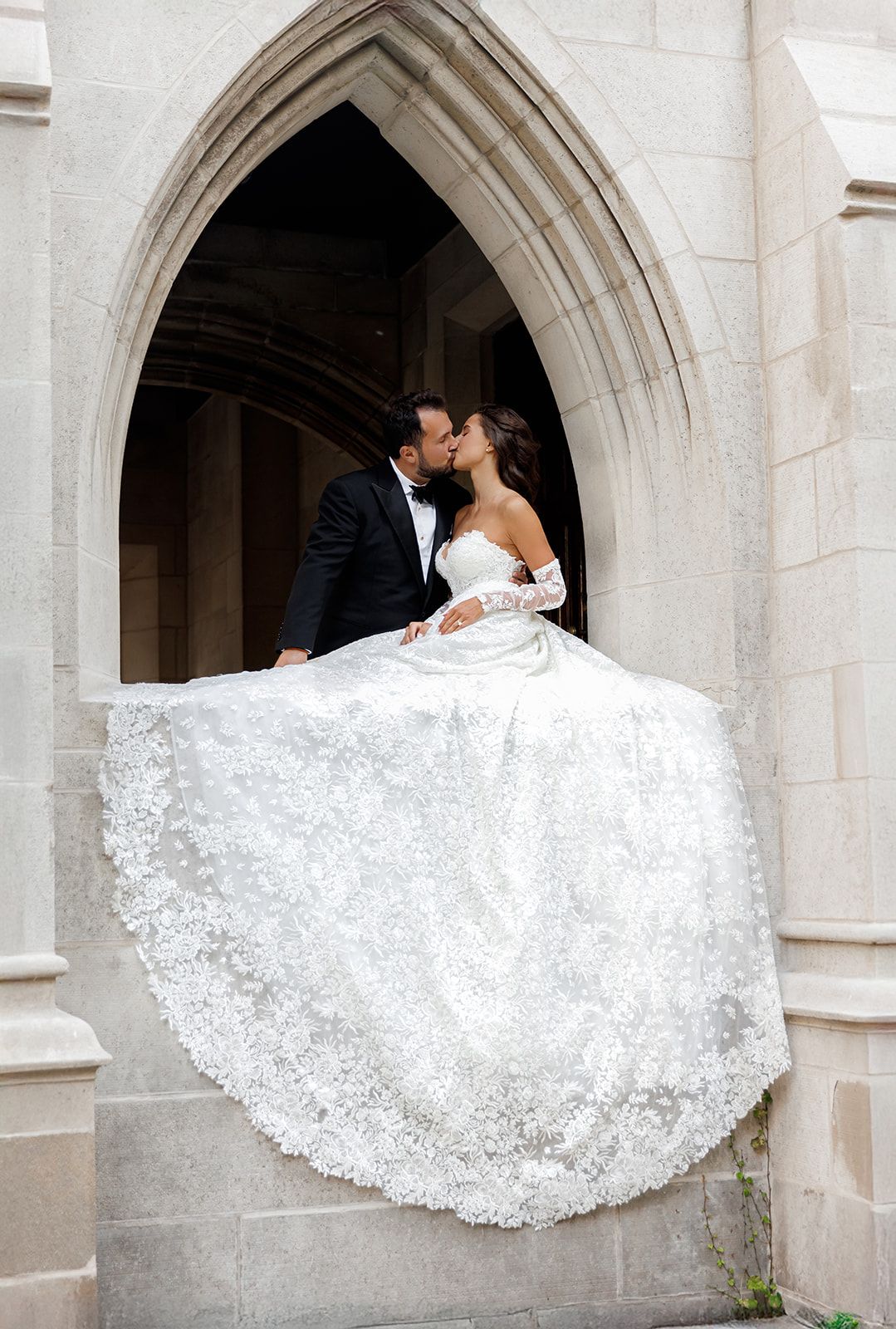 Bride and groom share a romantic kiss in a Gothic stone archway, her lace ballgown trailing elegantly below.