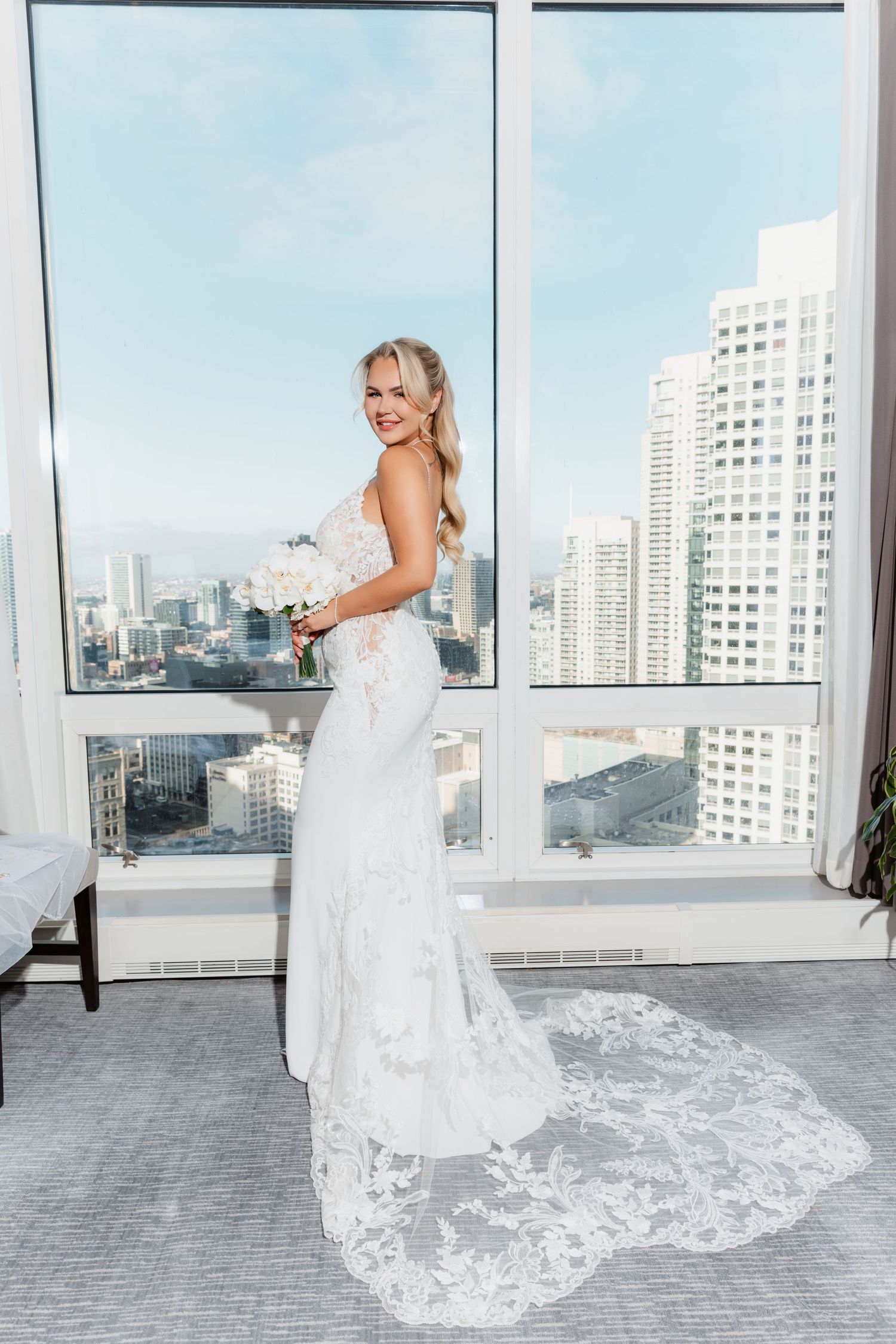 Bride in lace wedding gown with train holds white bouquet, posing before floor-to-ceiling windows with city skyline view.