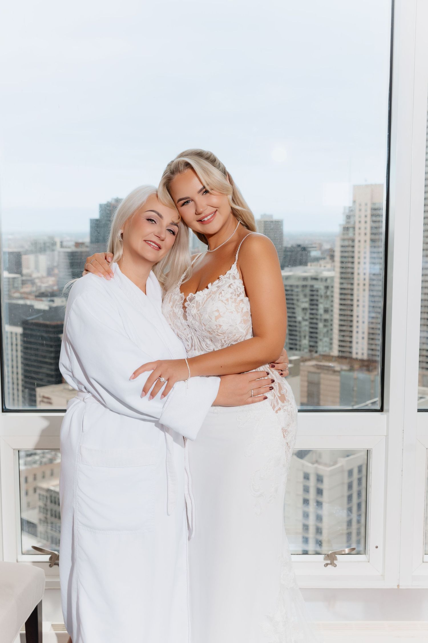 Bride in lace wedding gown embraces woman in white robe, smiling in front of city skyline window.