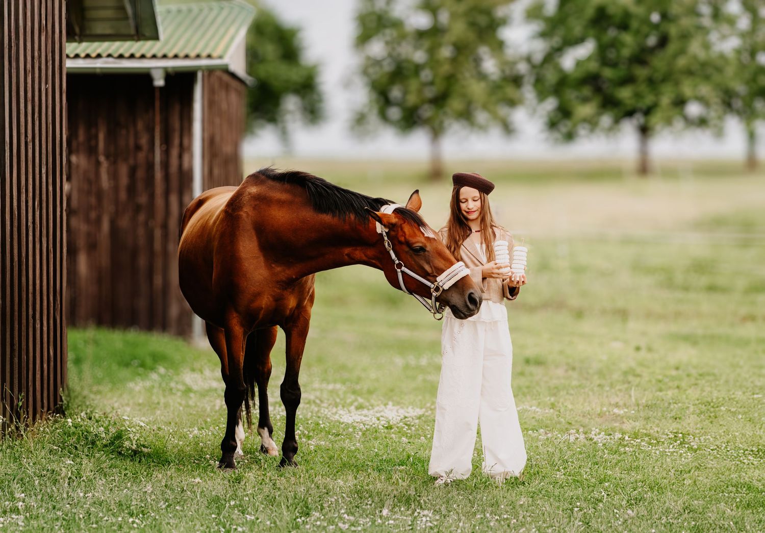Young woman in white dress holding books stands beside a brown horse on a green farm field near a wooden barn.