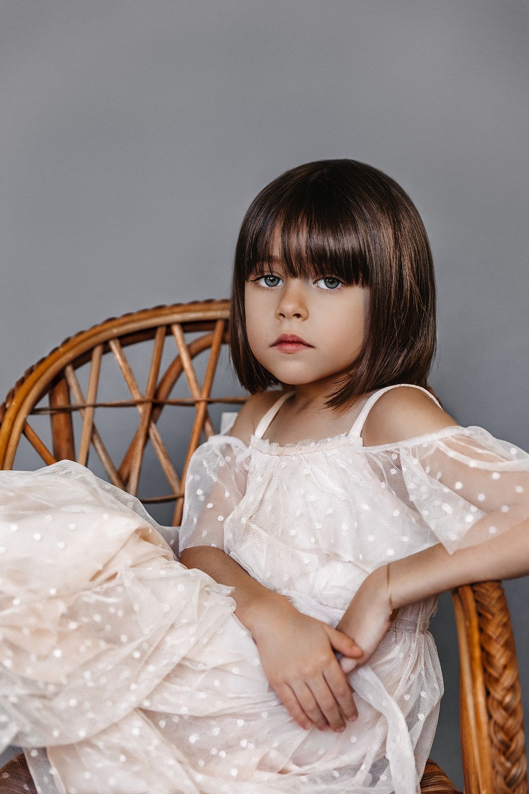A little girl with dark bob hair sits in a wicker chair wearing a white polka dot tulle dress on gray background.