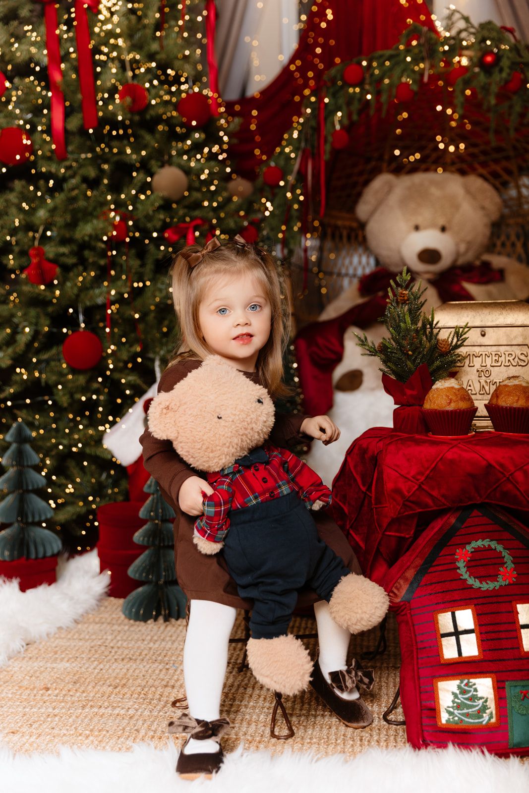 A toddler girl in a brown dress hugs a teddy bear beside a decorated Christmas tree with red ornaments and festive decor.