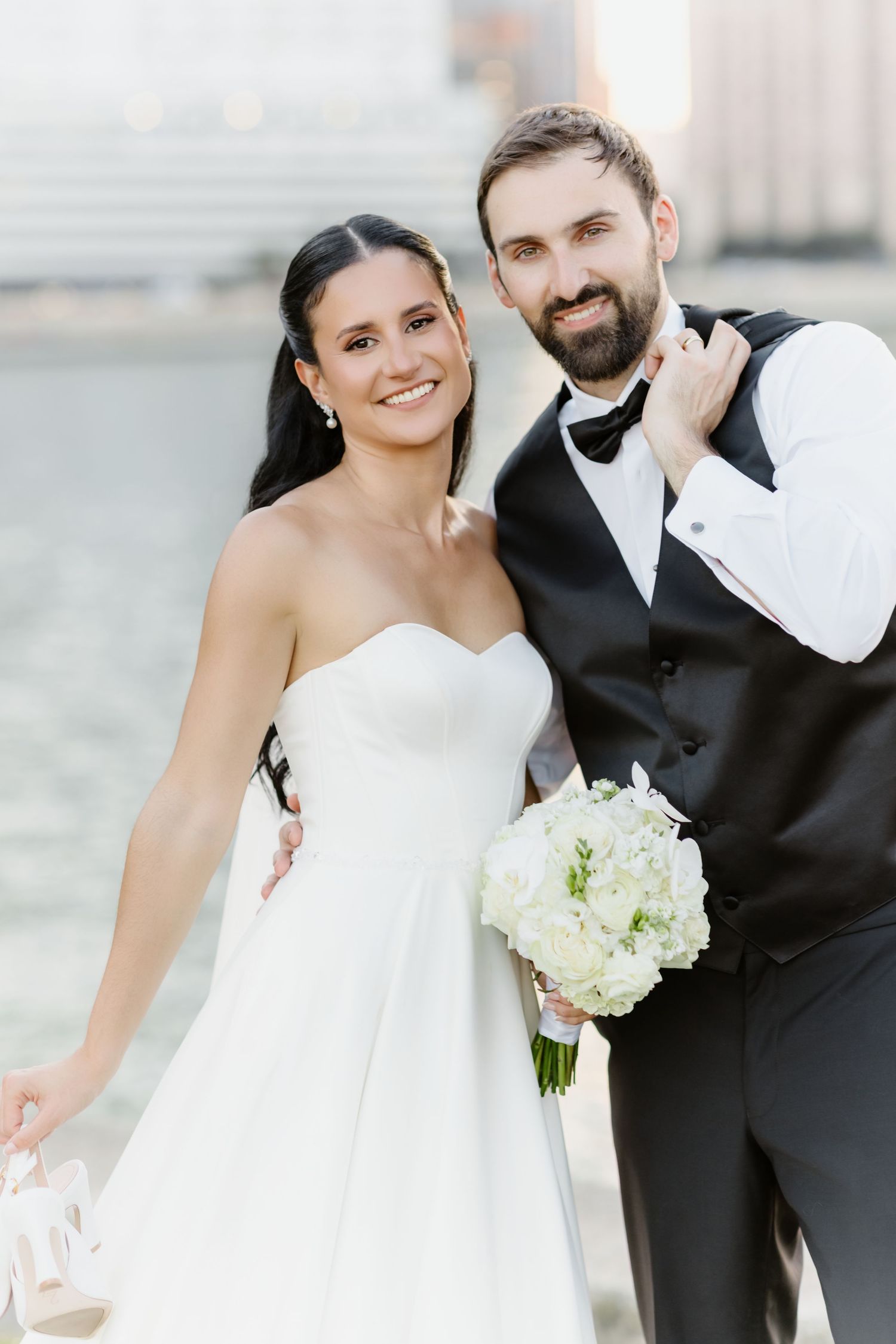 Smiling bride in strapless white gown and bearded groom in black tuxedo pose together holding white bouquet.