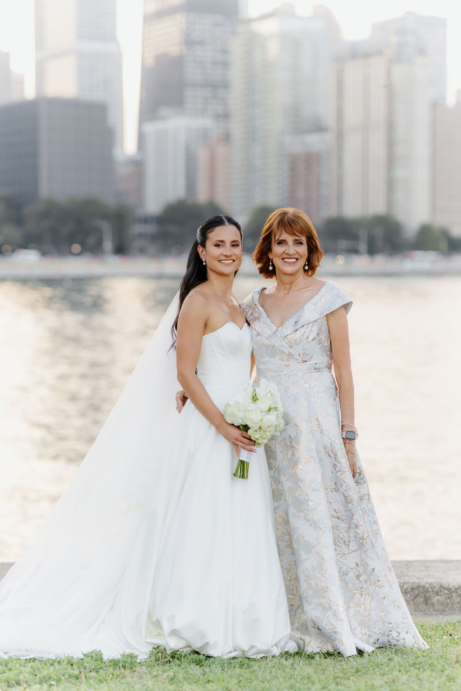 Bride in strapless white gown holds white bouquet posing with mother in silver dress against city skyline.