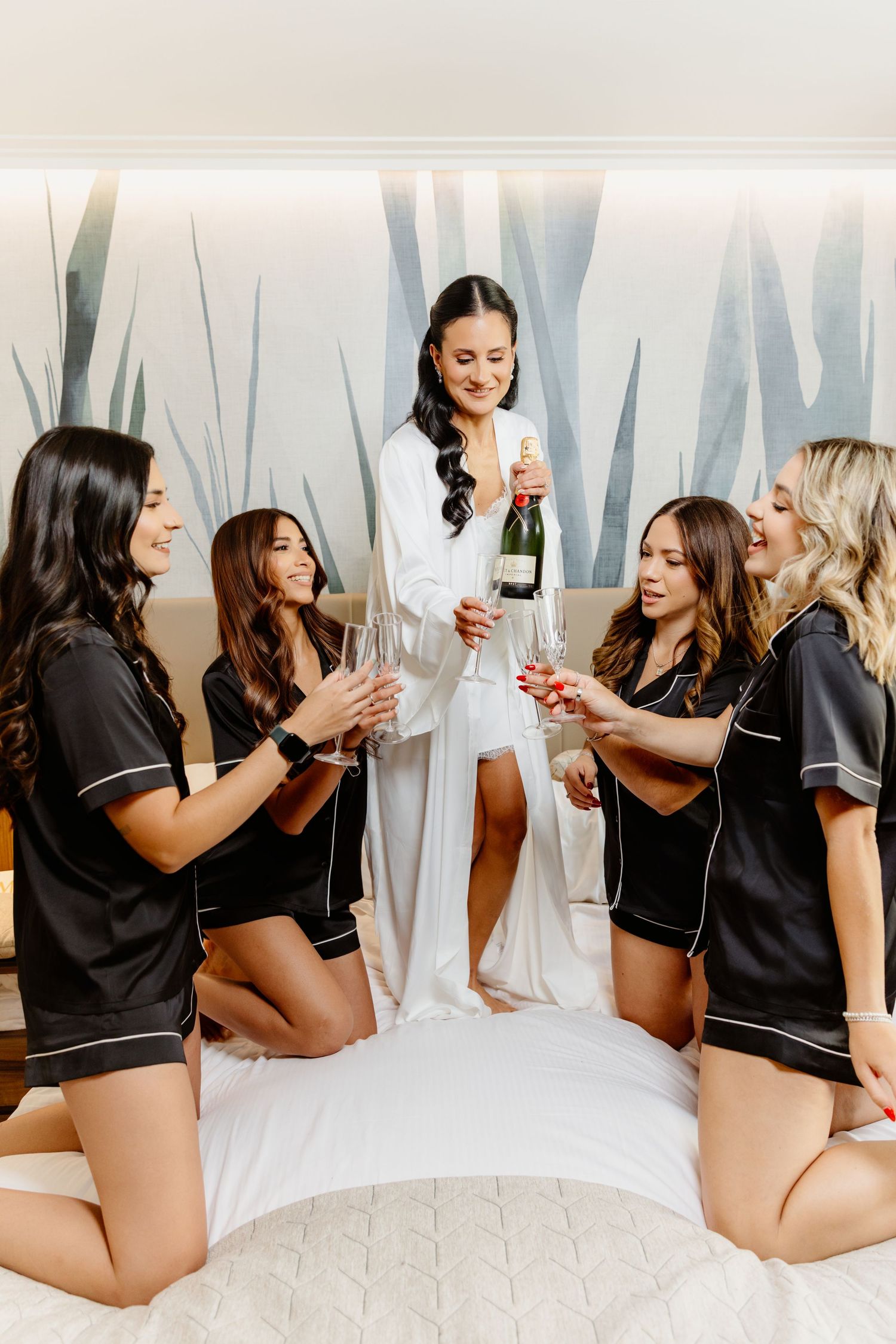 Bride in white robe pours champagne for four bridesmaids in black satin pajamas on hotel bed.