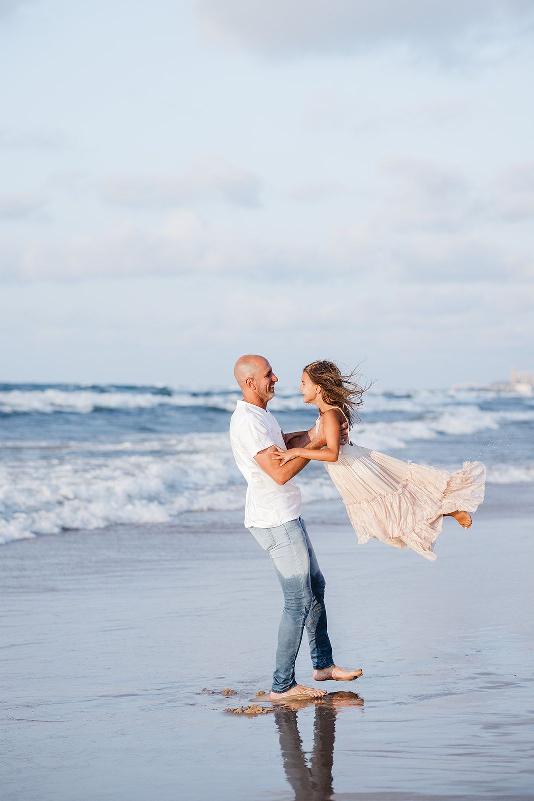 A couple shares a romantic moment on a wet beach, man lifting woman in flowing dress with ocean waves behind them.