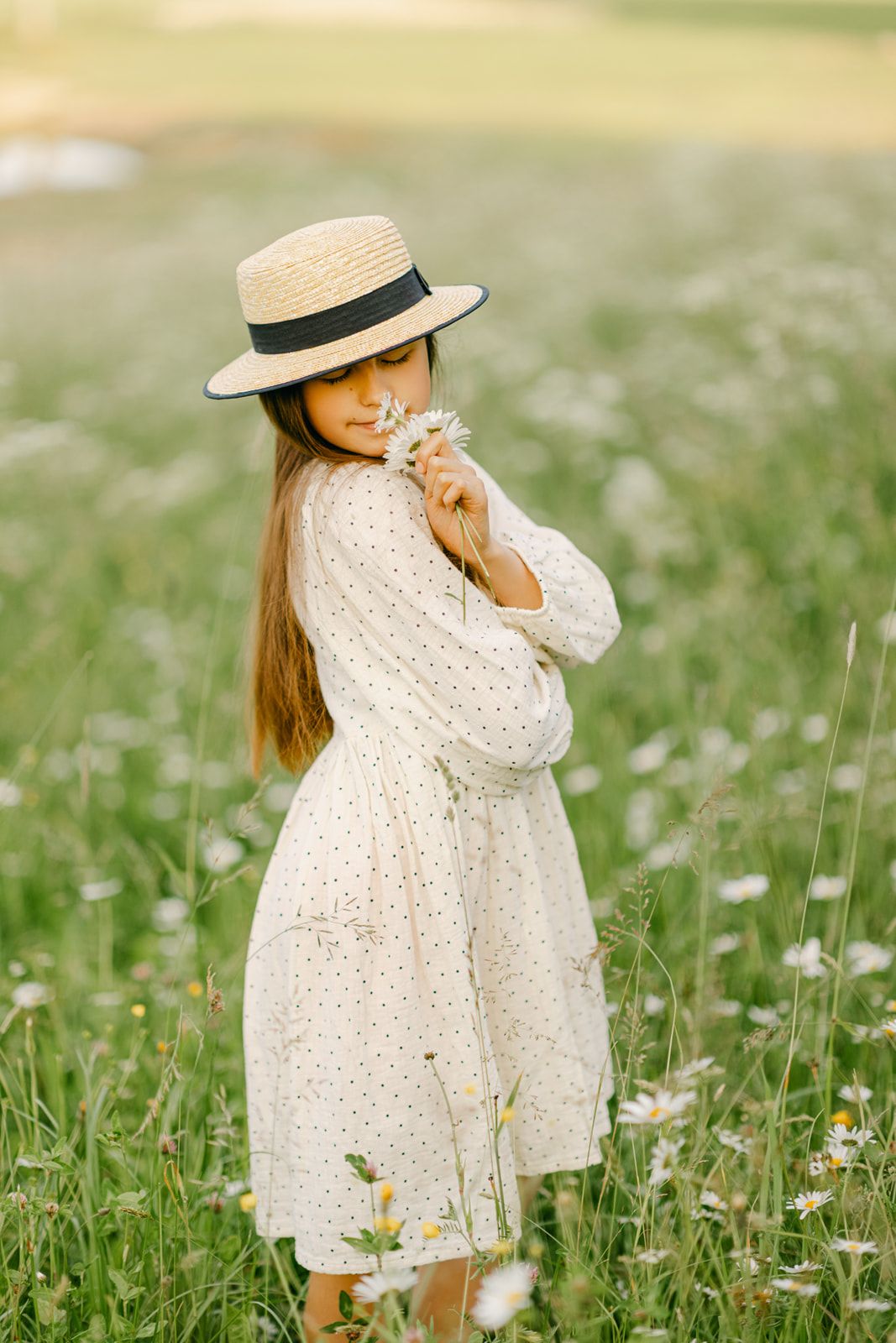Young girl in straw hat and white polka dot dress holds daisies in a blooming meadow.