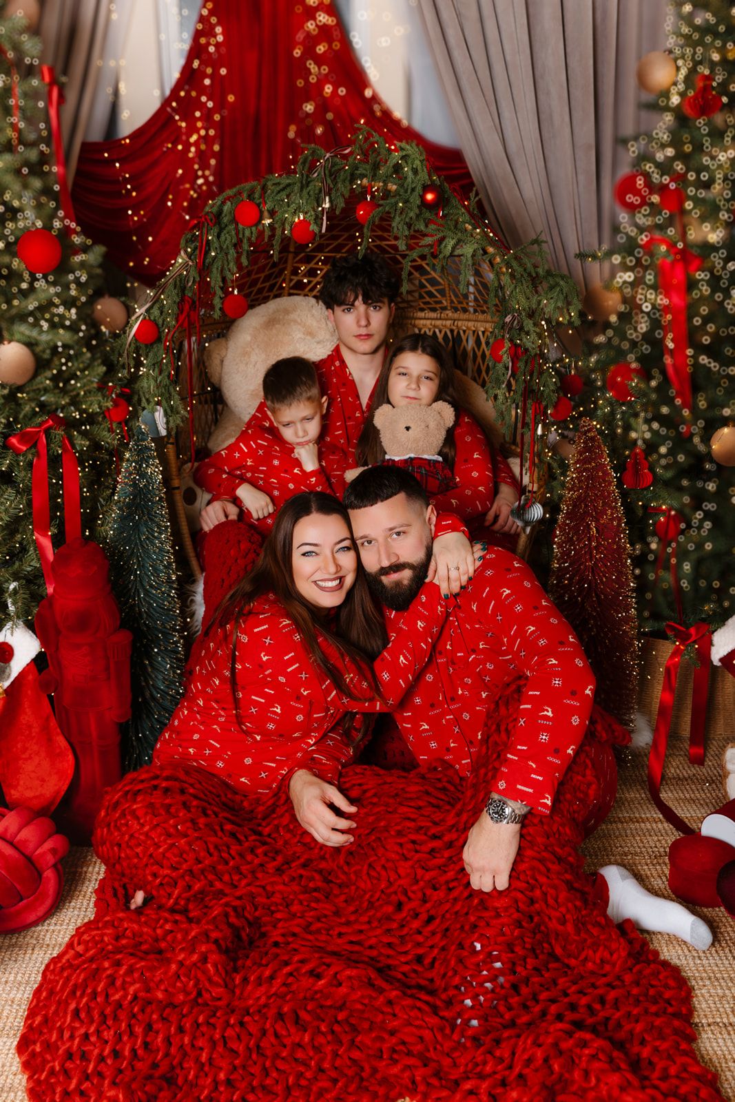 Family in matching red pajamas poses together among Christmas trees with festive decorations and holiday lights.