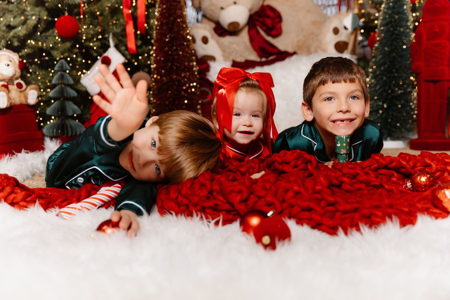 Three young children in green pajamas lying on white fur rug with red blanket near Christmas tree decorations.