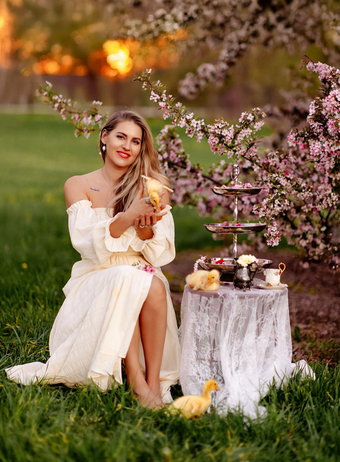 Woman in white off-shoulder dress sitting by blooming cherry tree with tiered dessert stand and ducklings at golden hour.