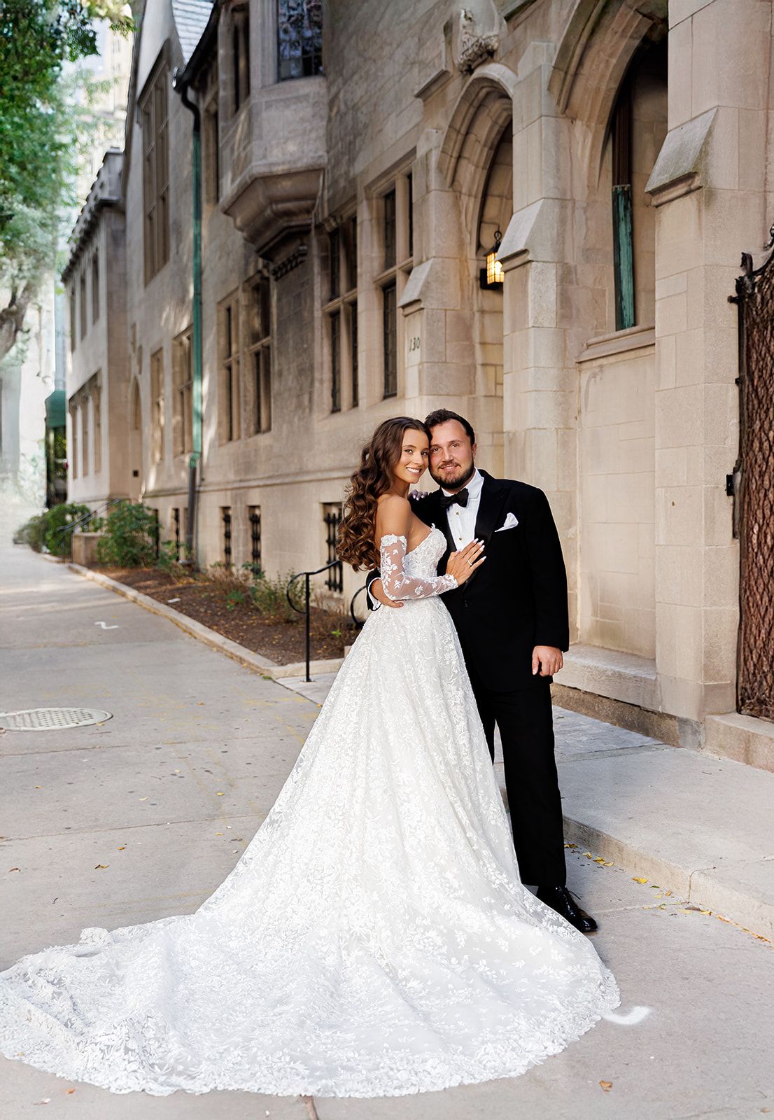 Bride in lace ball gown with groom in tuxedo posing on urban sidewalk beside historic stone building.