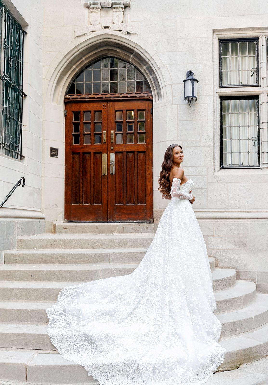 Bride in a stunning lace off-shoulder wedding gown with a long train poses on stone steps before ornate wooden doors.