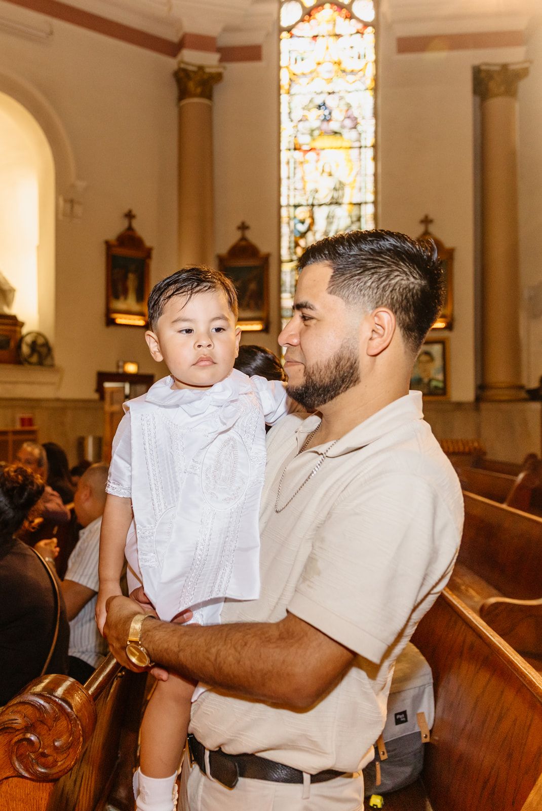 A man holds a young boy dressed in white at a church baptism ceremony with stained glass windows.
