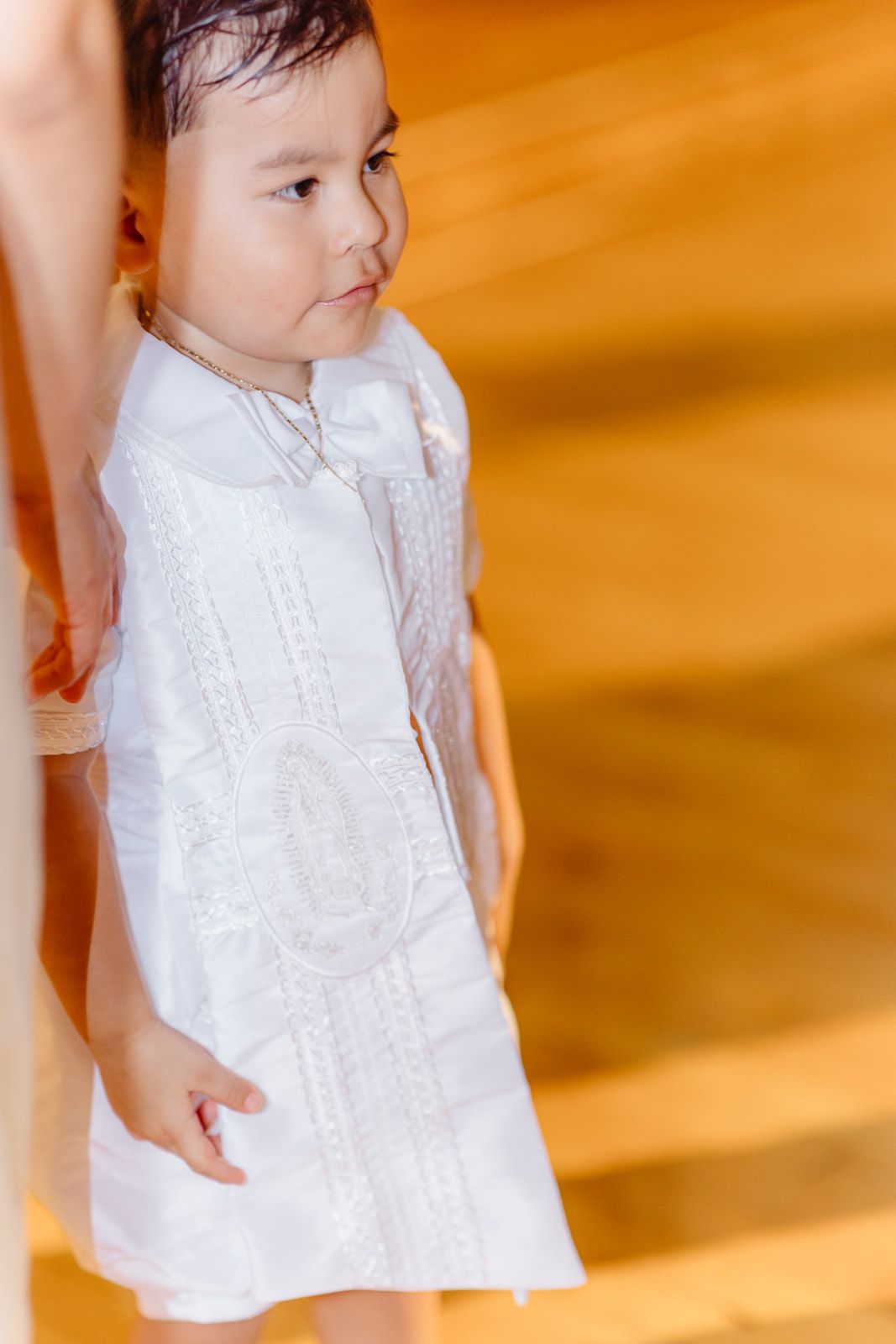 A toddler in a white embroidered Barong Tagalog dress stands in a warmly lit church during a baptism ceremony.