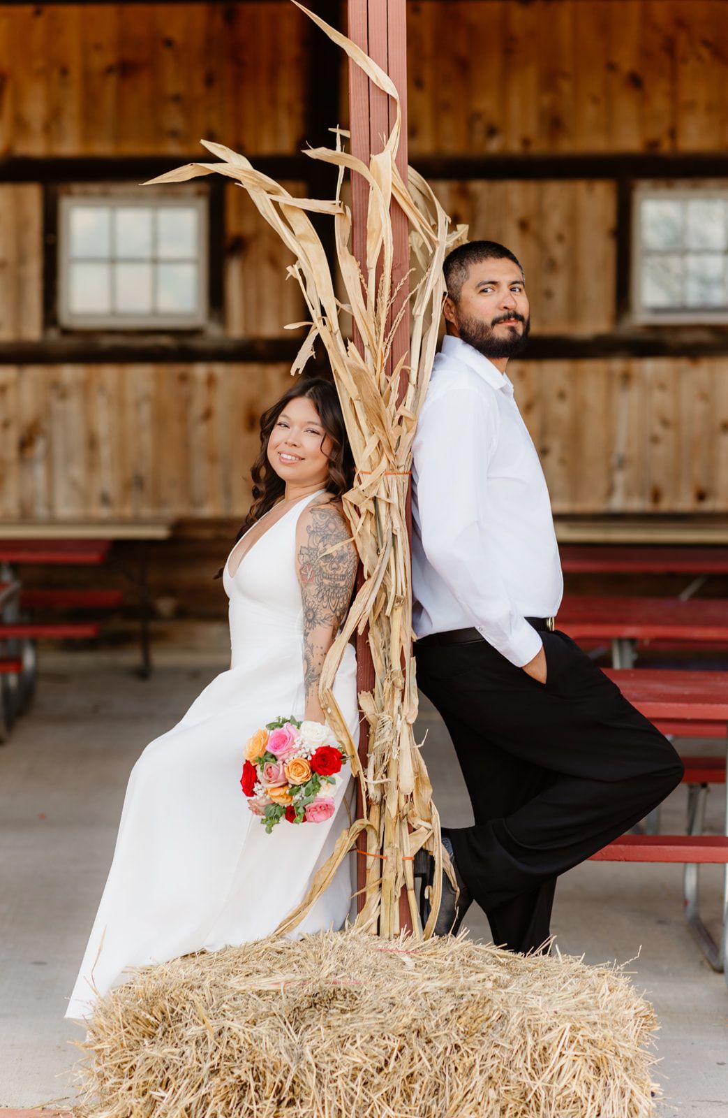Bride and groom pose back-to-back beside dried corn stalks on hay bale in rustic wooden barn wedding venue.