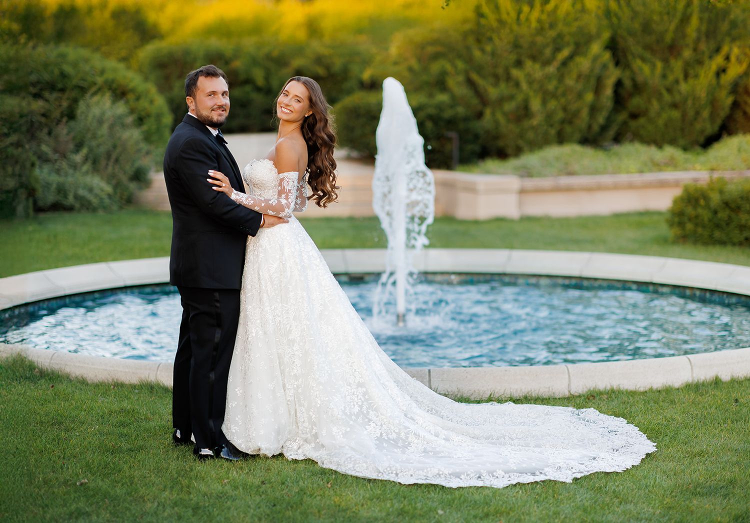 Couple sharing a romantic moment by a garden fountain, bride in a flowing lace gown with a long train.