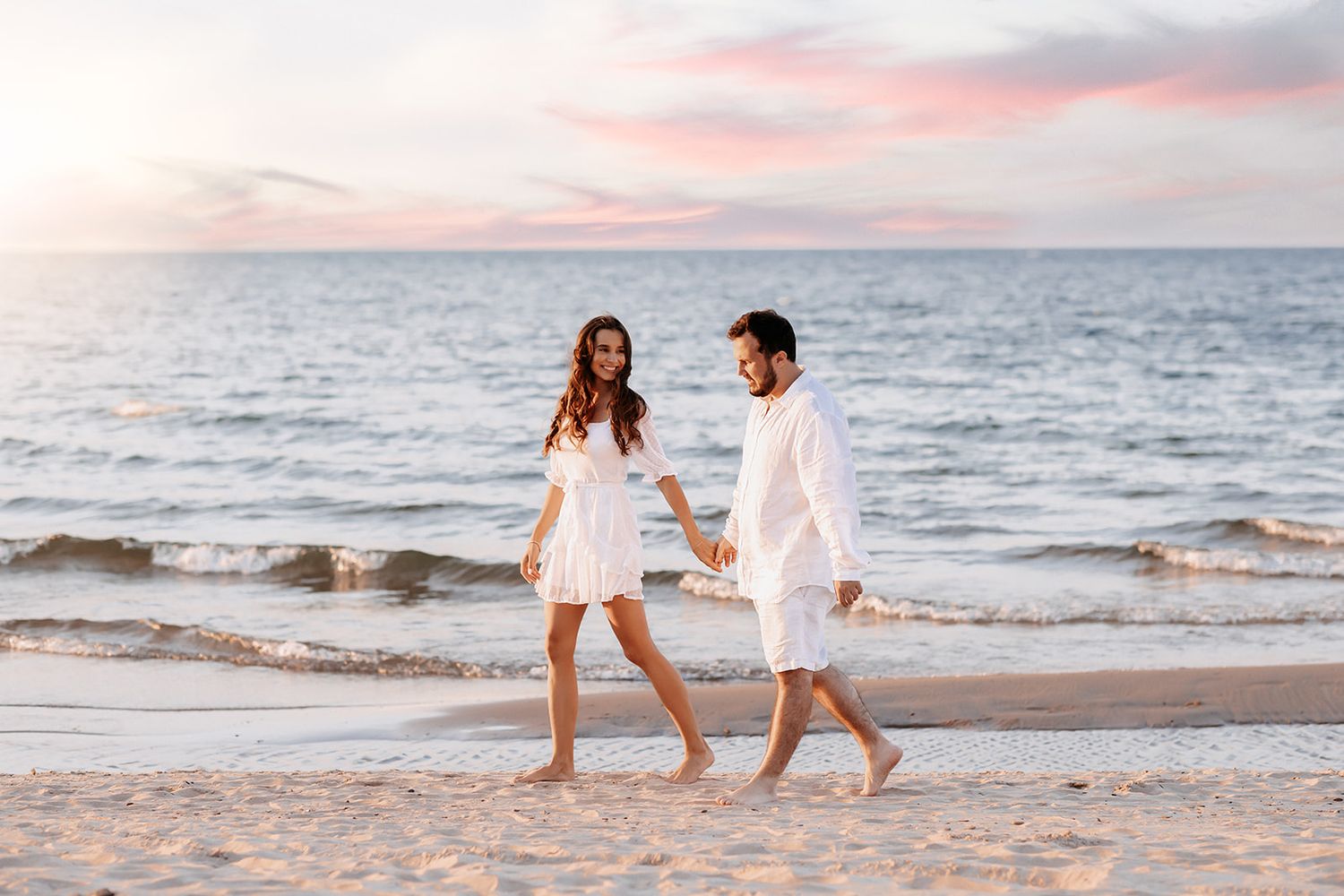 Happy couple in white outfits holding hands and walking along the shoreline at sunset.