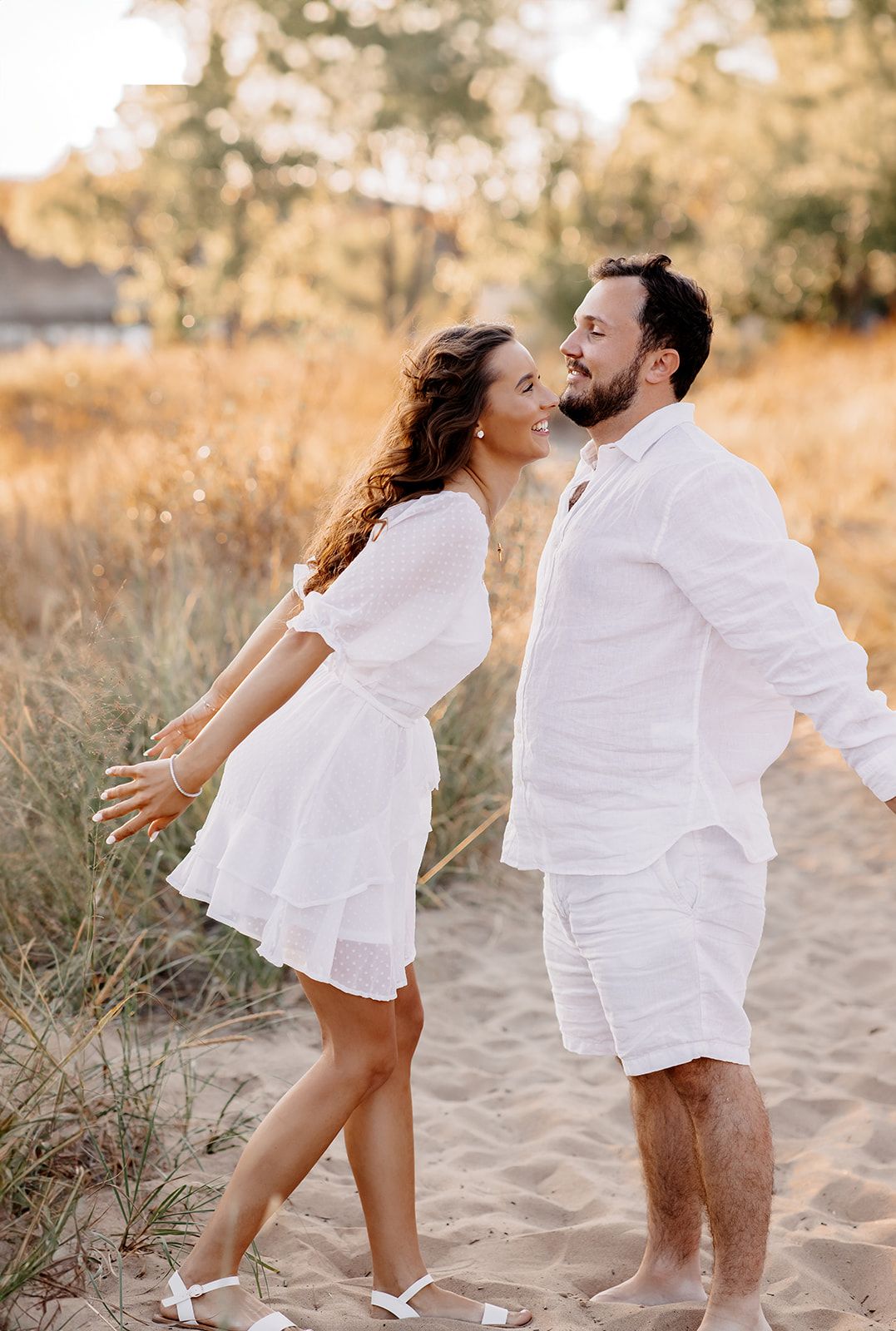Couple in matching white outfits dancing playfully on sandy beach path with golden grass backdrop.