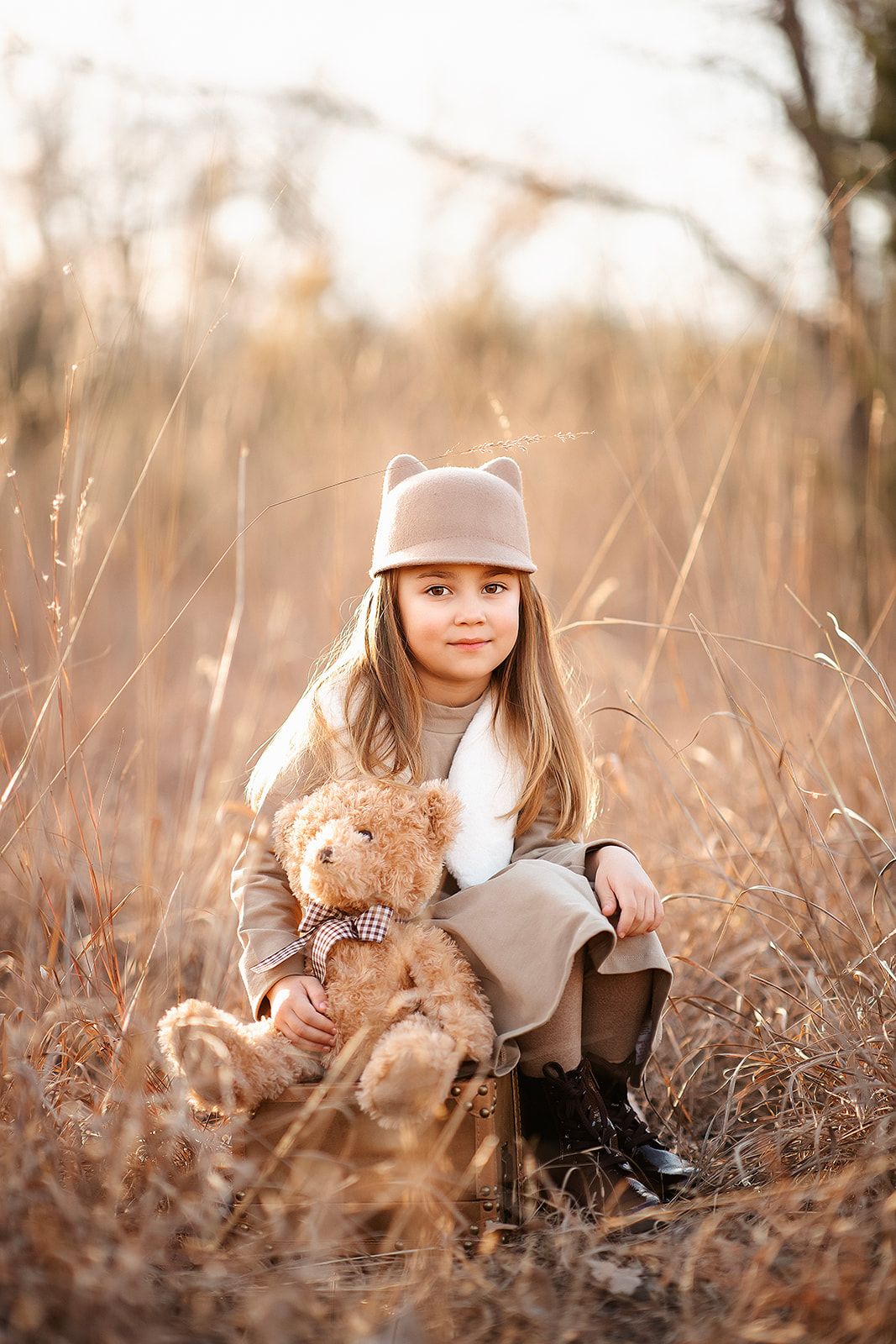 Young girl in beige coat and bear-ear hat sits in golden field holding a teddy bear with plaid bow tie.