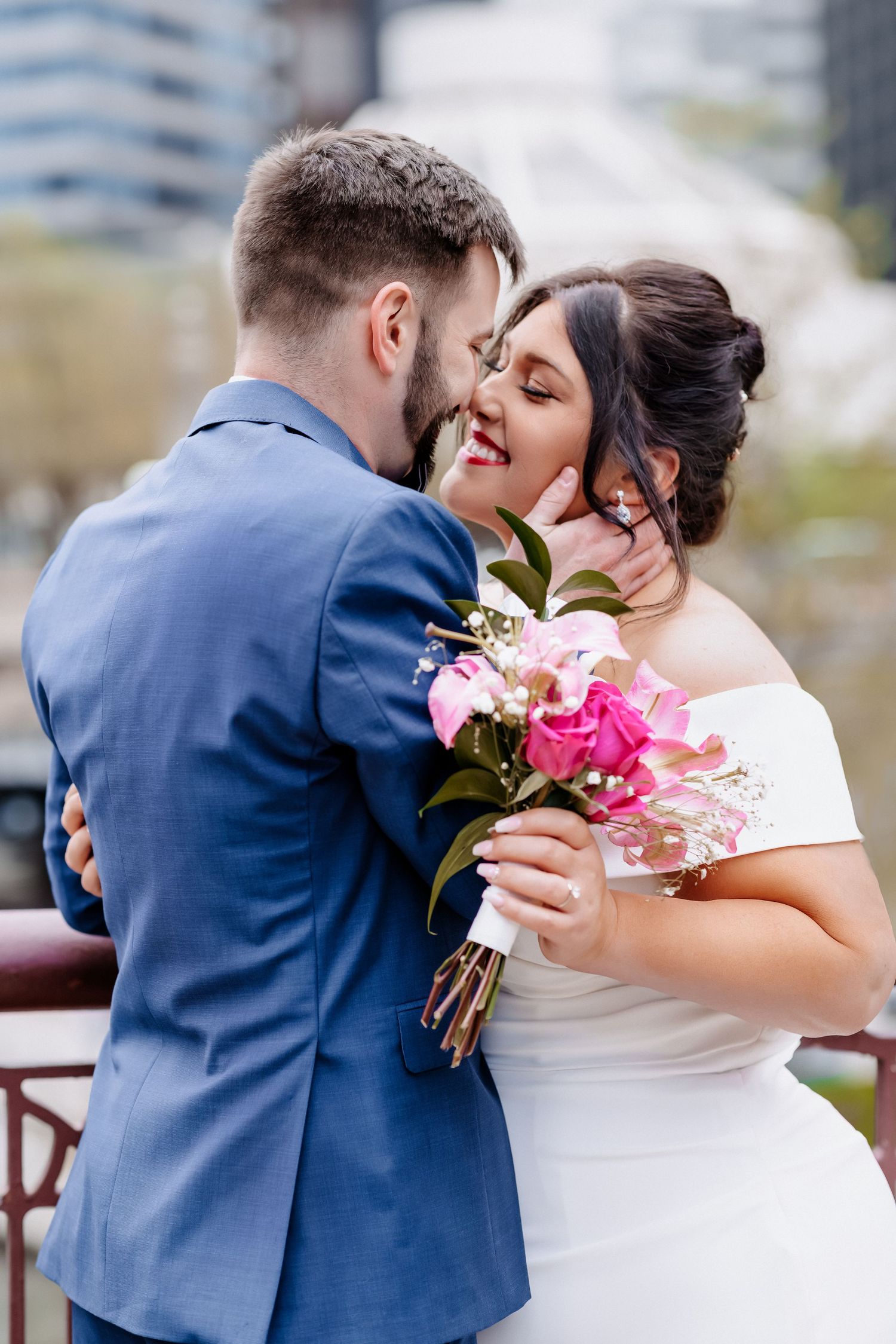 Groom in blue suit kisses bride in white off-shoulder gown holding pink floral bouquet on urban bridge.