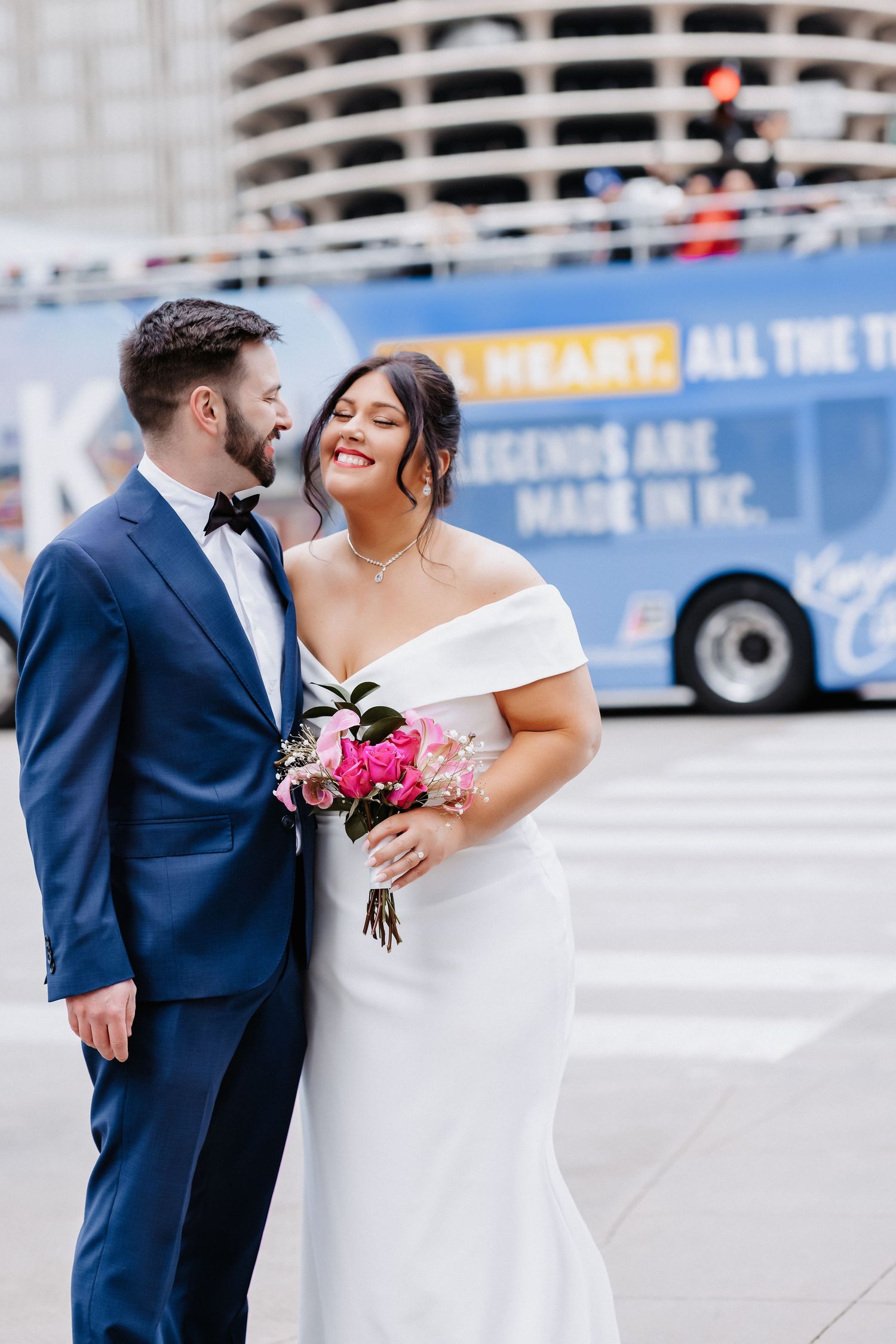 Elegant wedding couple embraces on Chicago street, bride in off-shoulder gown with pink bouquet, groom in navy suit.