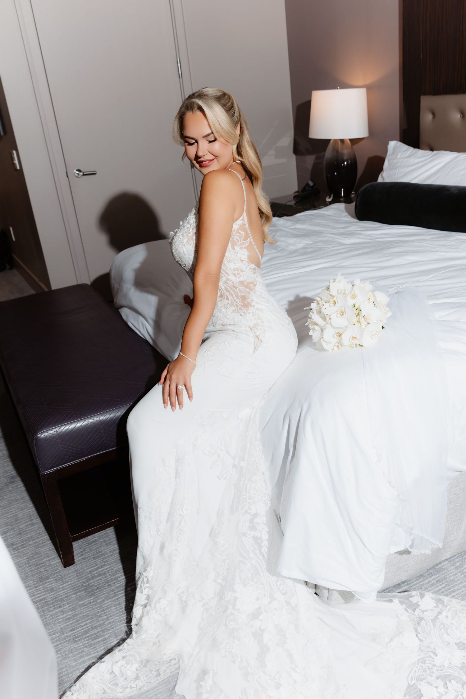 Radiant blonde bride in backless lace wedding gown with long train smiles beside white bouquet on hotel bed.
