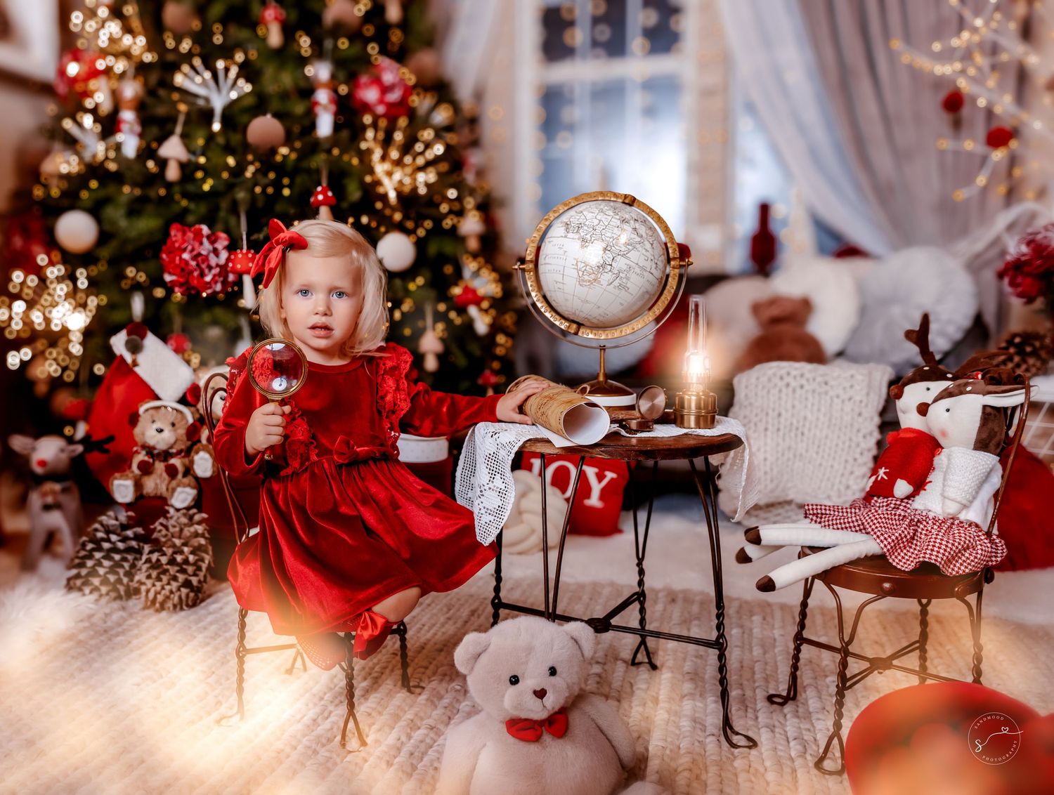 Toddler girl in red dress sits near Christmas tree with teddy bears, globe, and candle decorations.