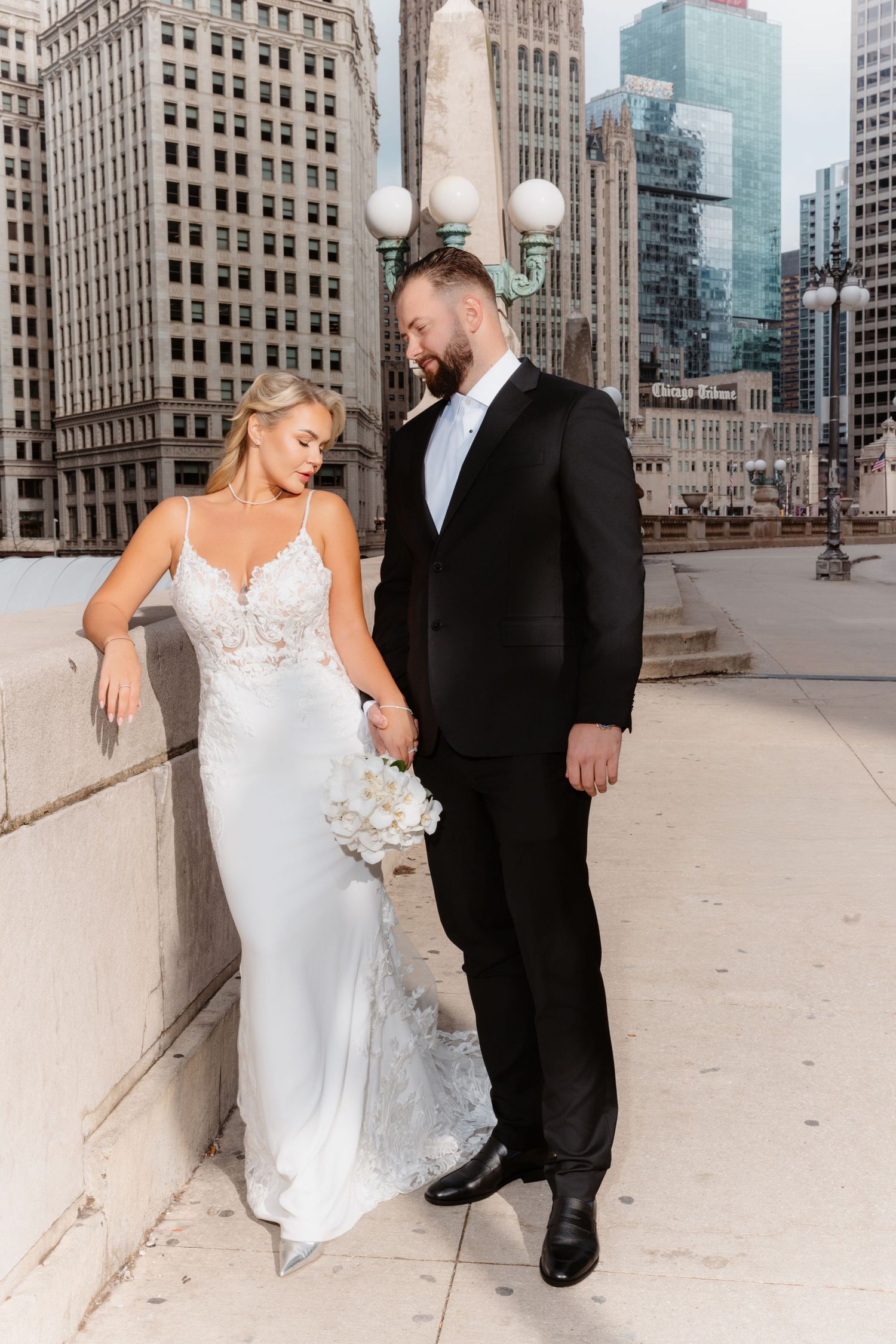 Groom in black tuxedo holds brides hand as she leans against stone wall on Chicago bridge.