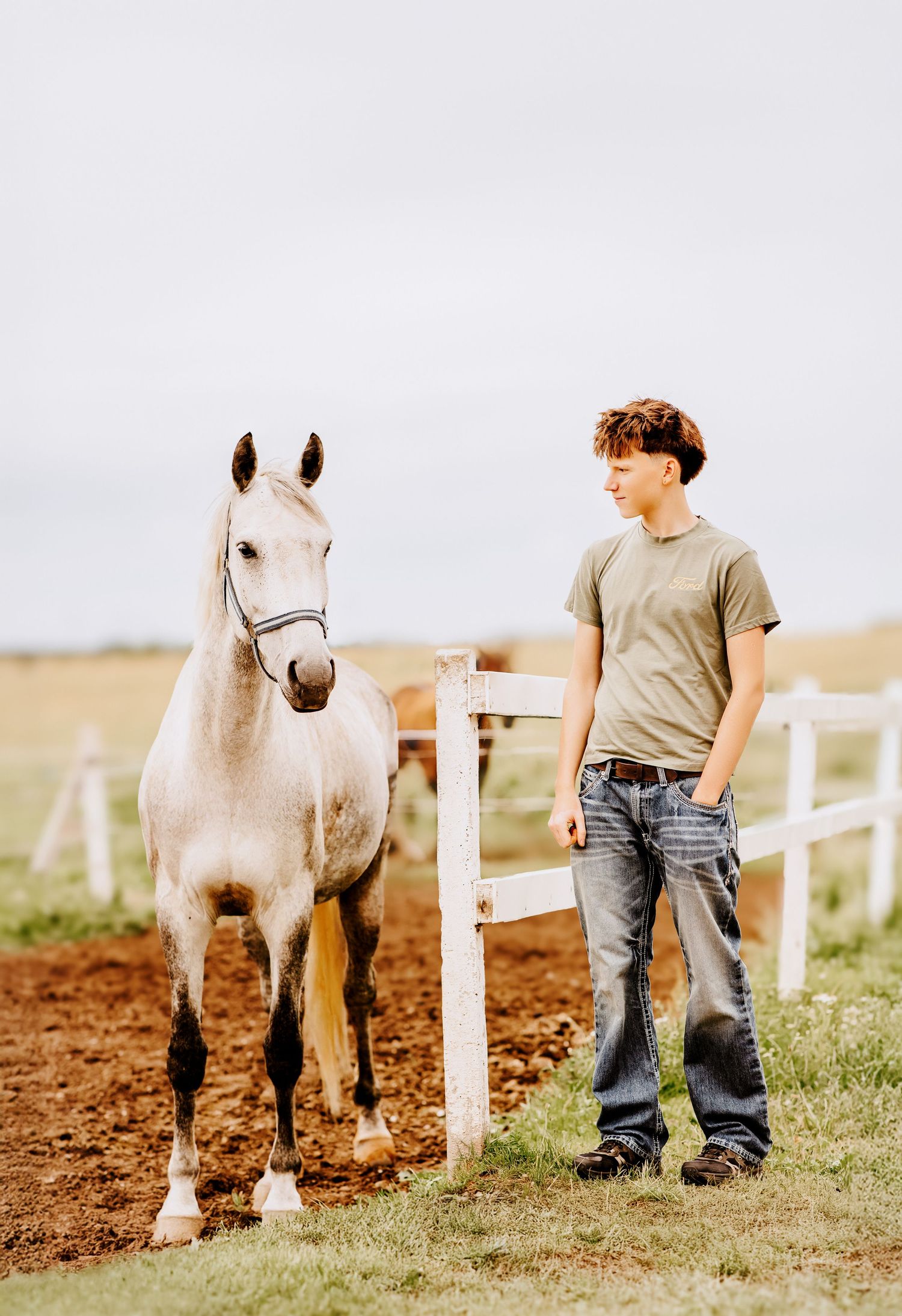 A teenage boy in jeans and a gray t-shirt stands beside a white horse near a white fence on a farm.