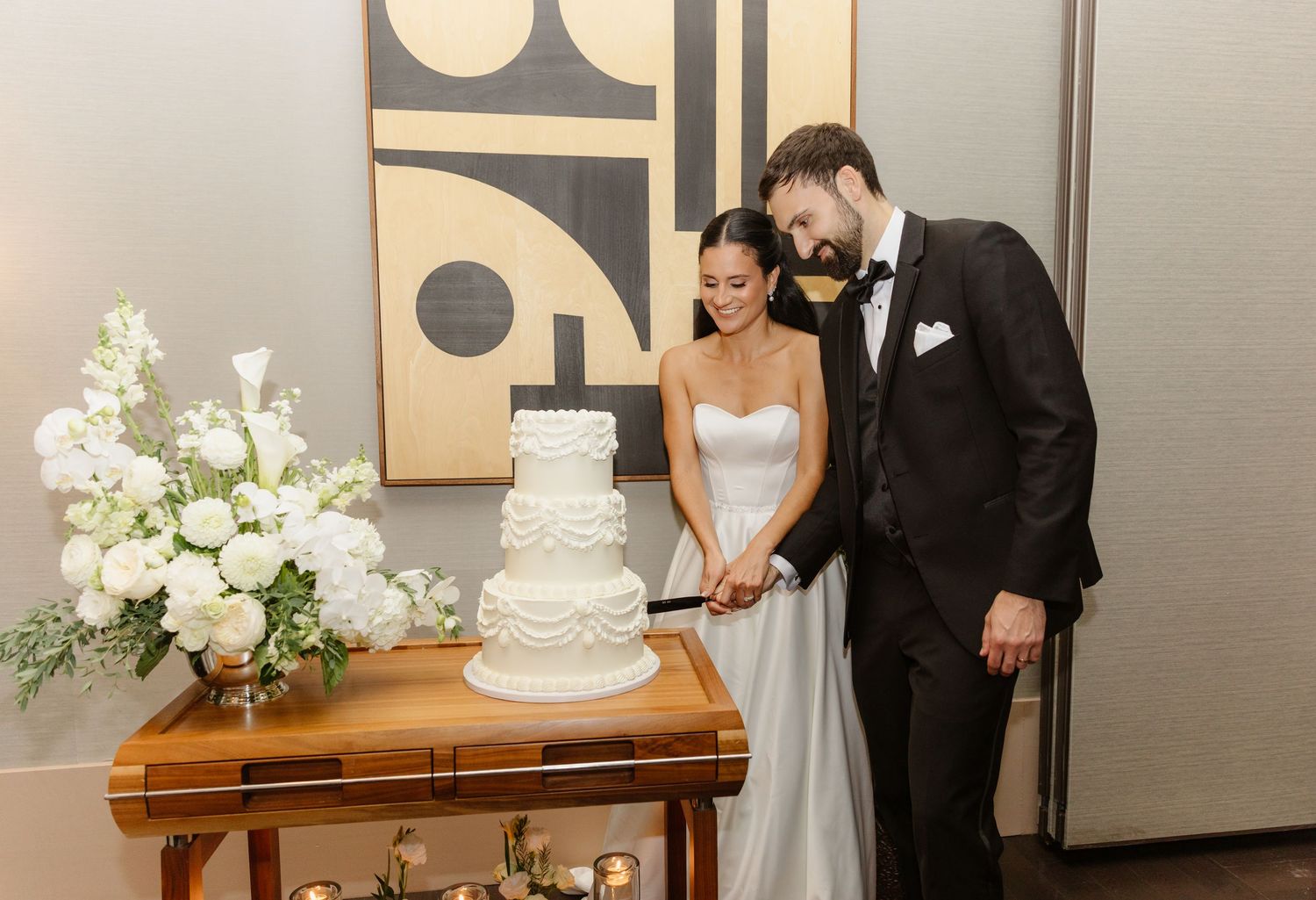 Bride and groom cutting a white multi-tiered wedding cake at their reception beside a lush white floral display.
