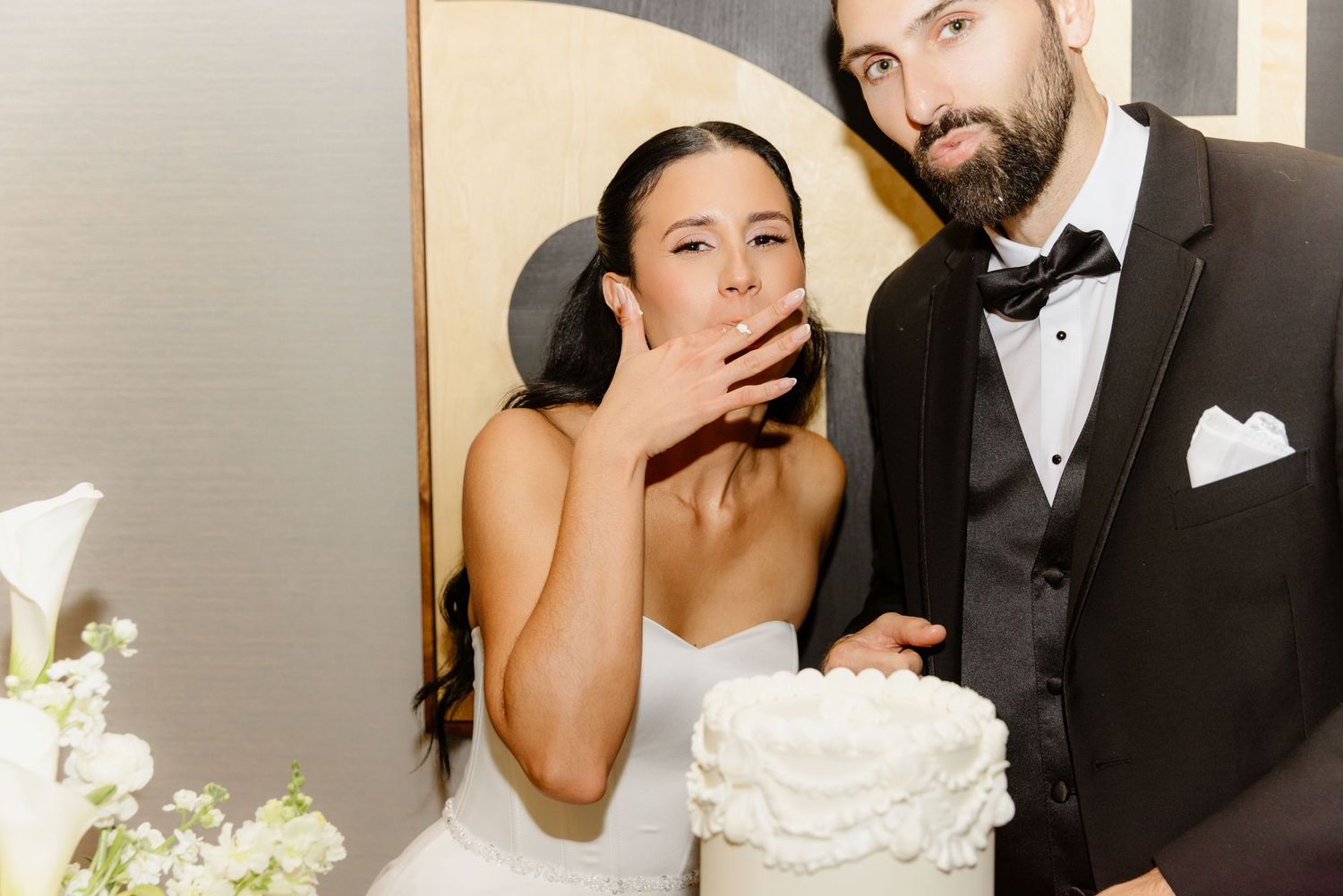 Bride tasting wedding cake while groom smiles beside her at elegant white tiered cake cutting ceremony.