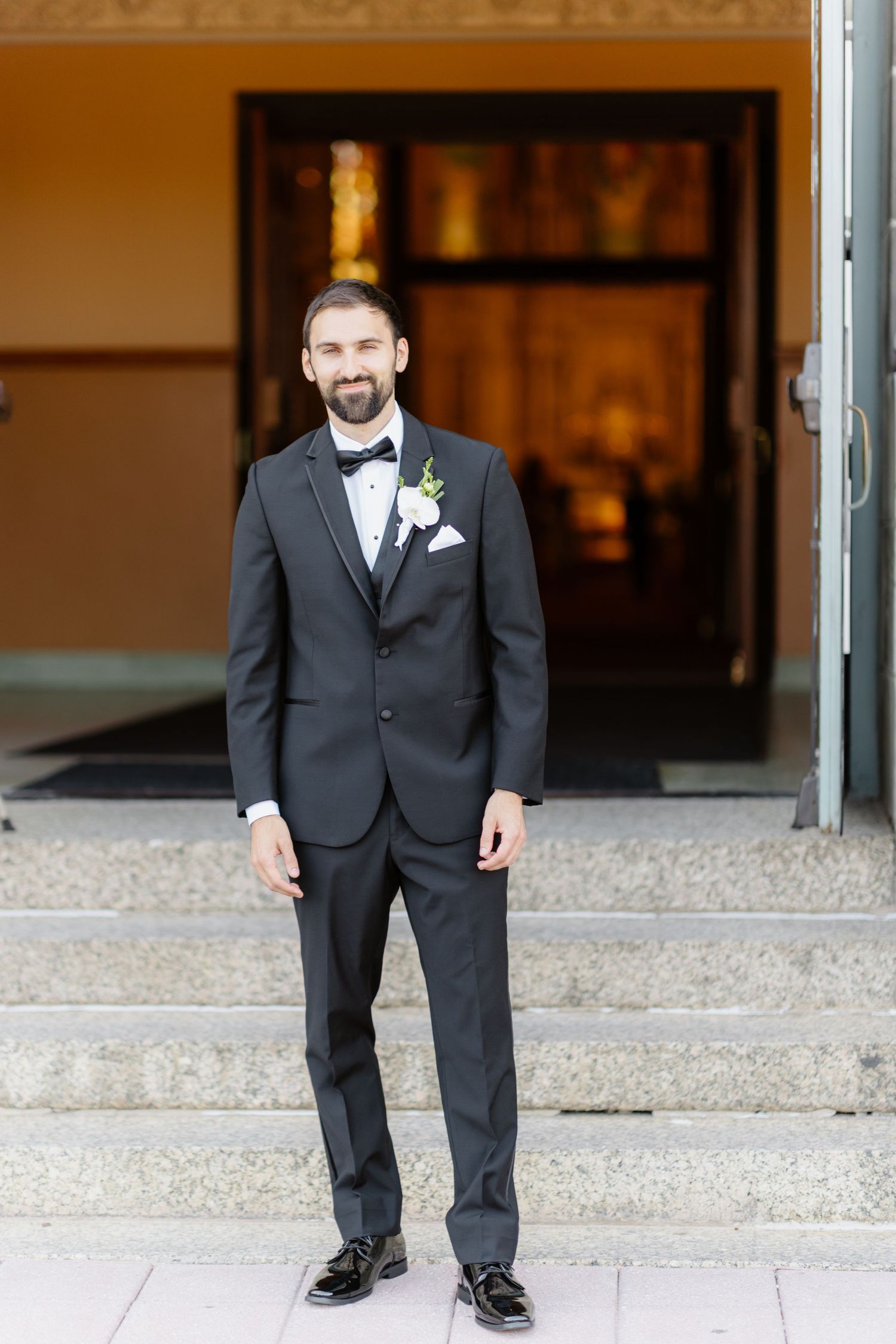 Dapper bearded groom in black tuxedo standing on church steps with white floral boutonniere.