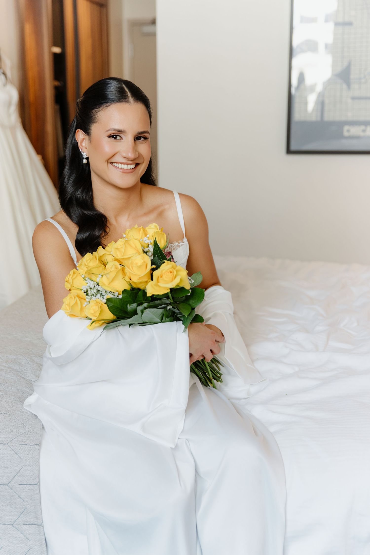 Radiant bride in white satin robe holds vibrant yellow rose bouquet while sitting on white bed in bright hotel room.