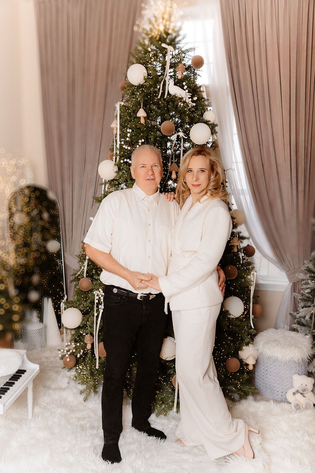 Elegant couple in white outfits posing together in front of a beautifully decorated Christmas tree.