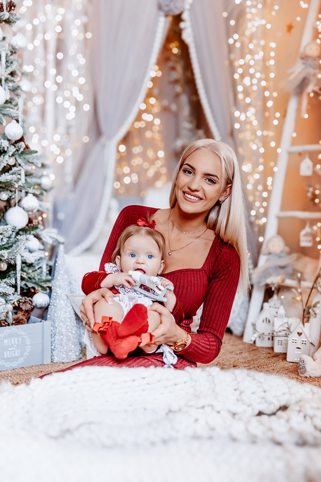 Smiling blonde mother in red dress holds baby girl with bow in festive Christmas decorated room with lights.