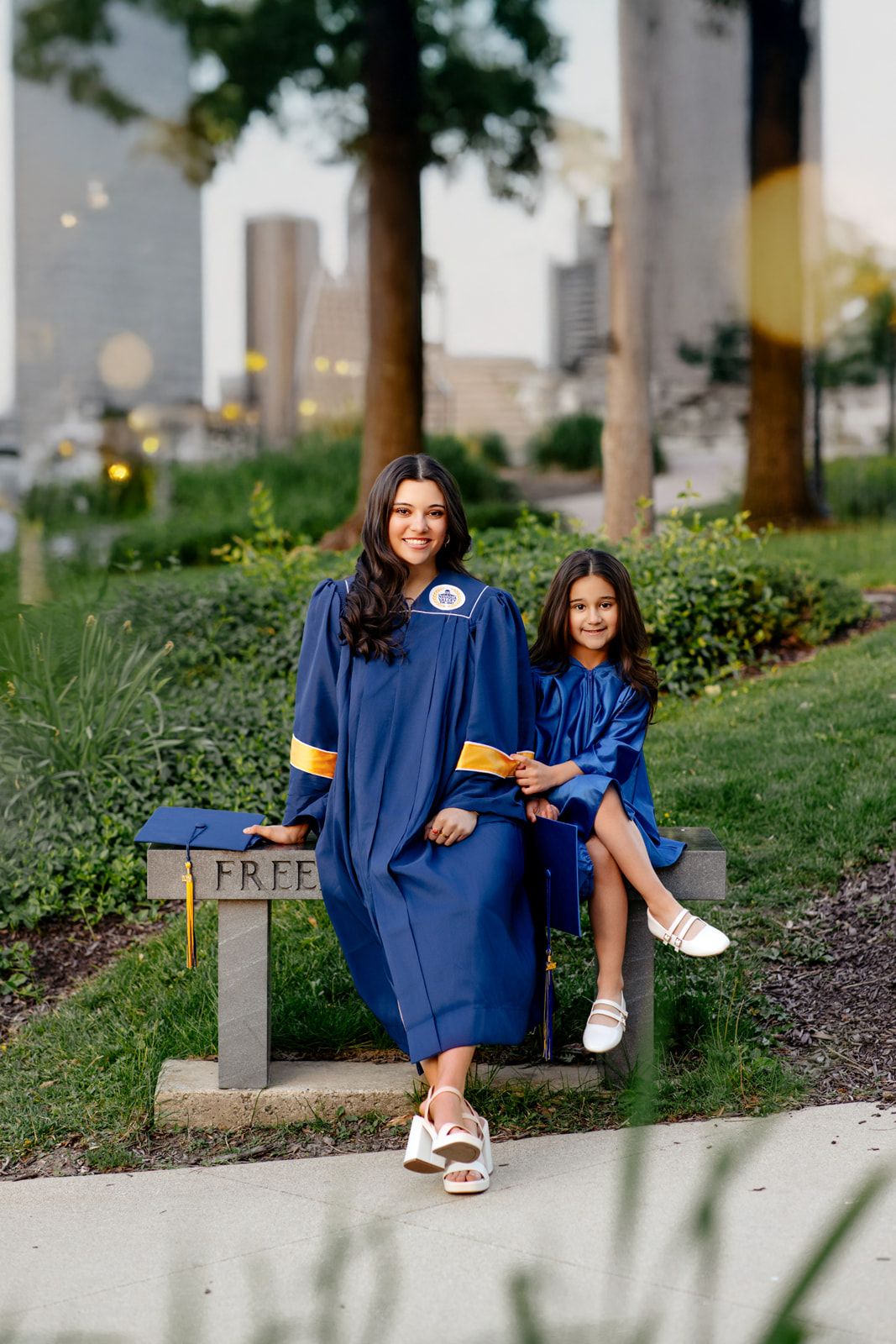 Graduate in blue gown posing with a young girl in matching gown on an outdoor bench.