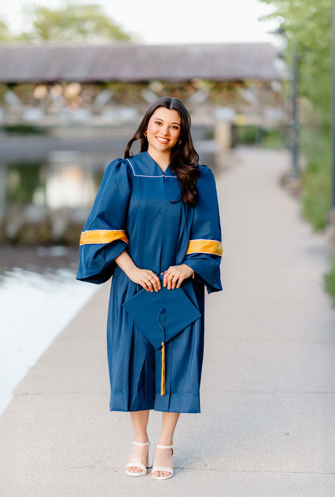 Young woman in navy graduation gown holding cap while posing on a scenic waterfront path.