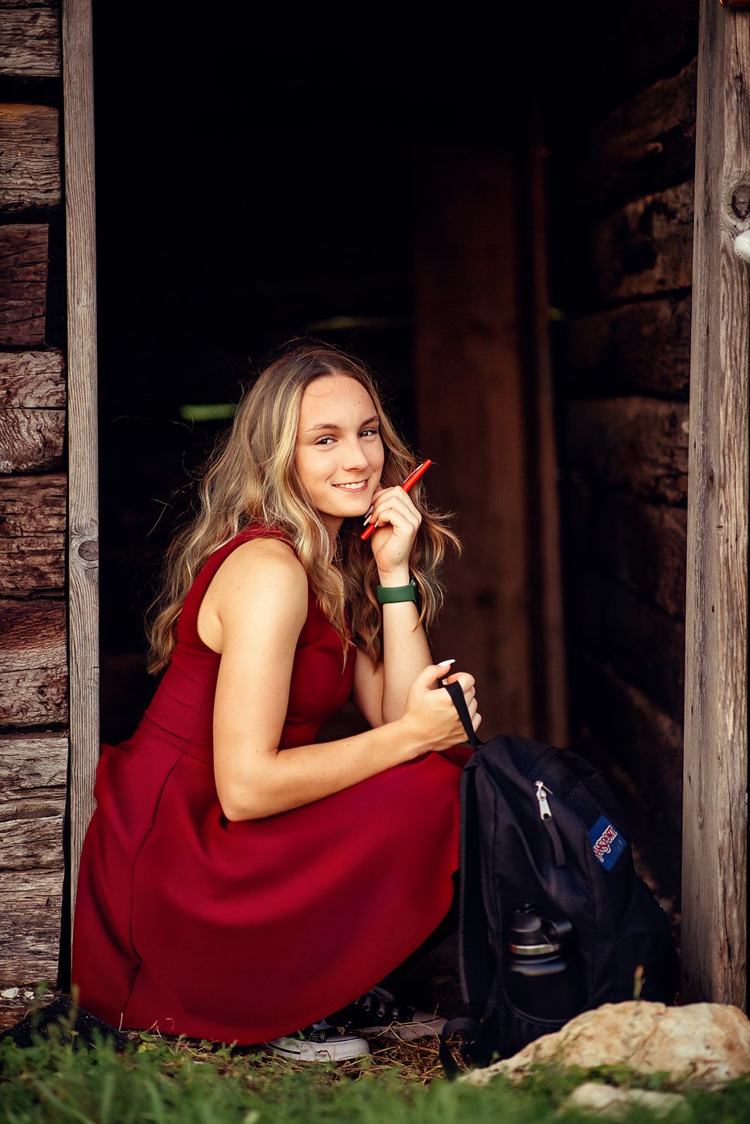 Smiling blonde girl in red dress crouching in rustic doorway holding a red pencil and backpack.